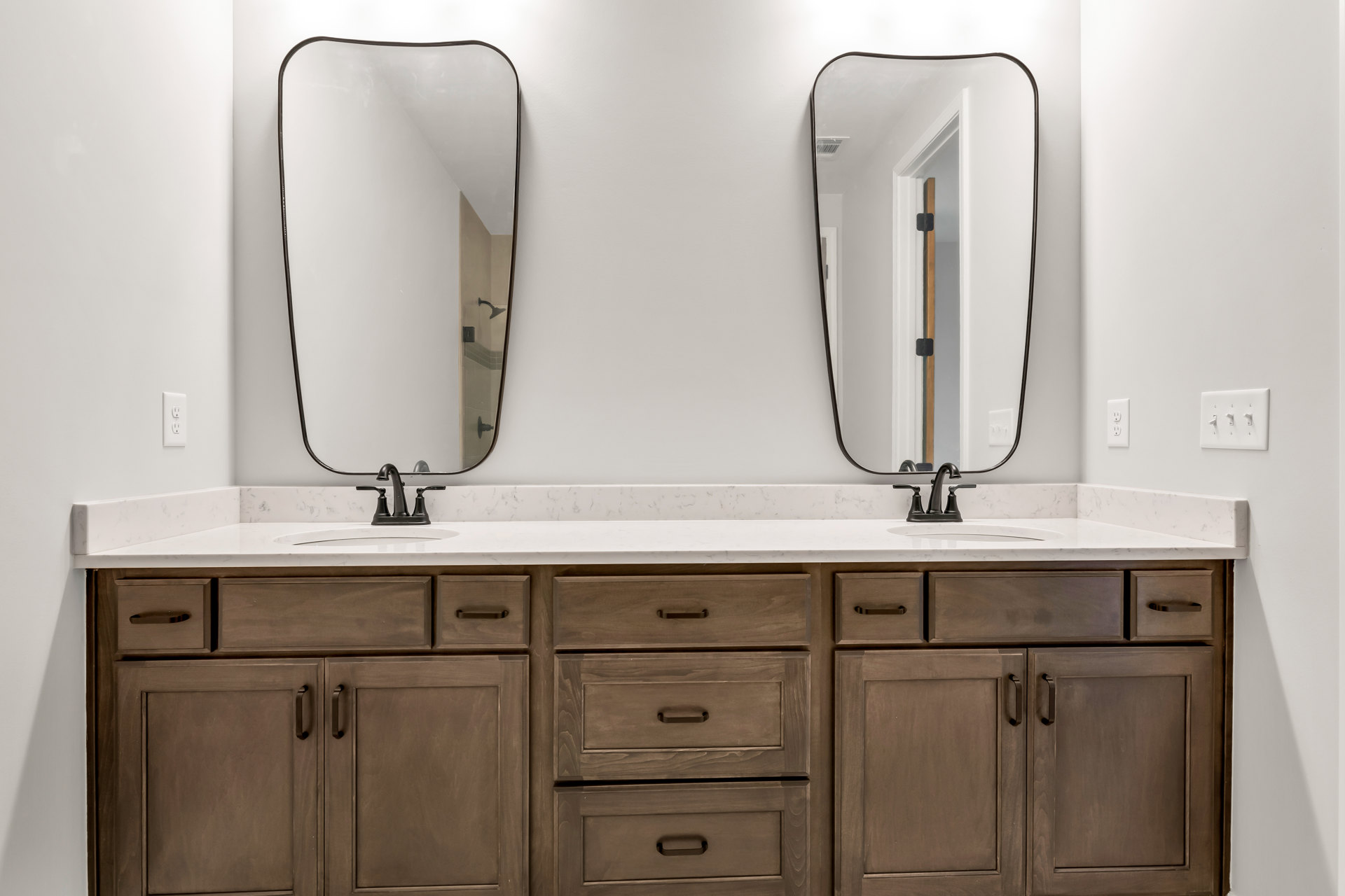 Double vanity bathroom with two undermount sinks, wide wall mirror, white quartz countertop, shaker-style drawers and cabinets, chrome faucets, and a light switch on the wall.