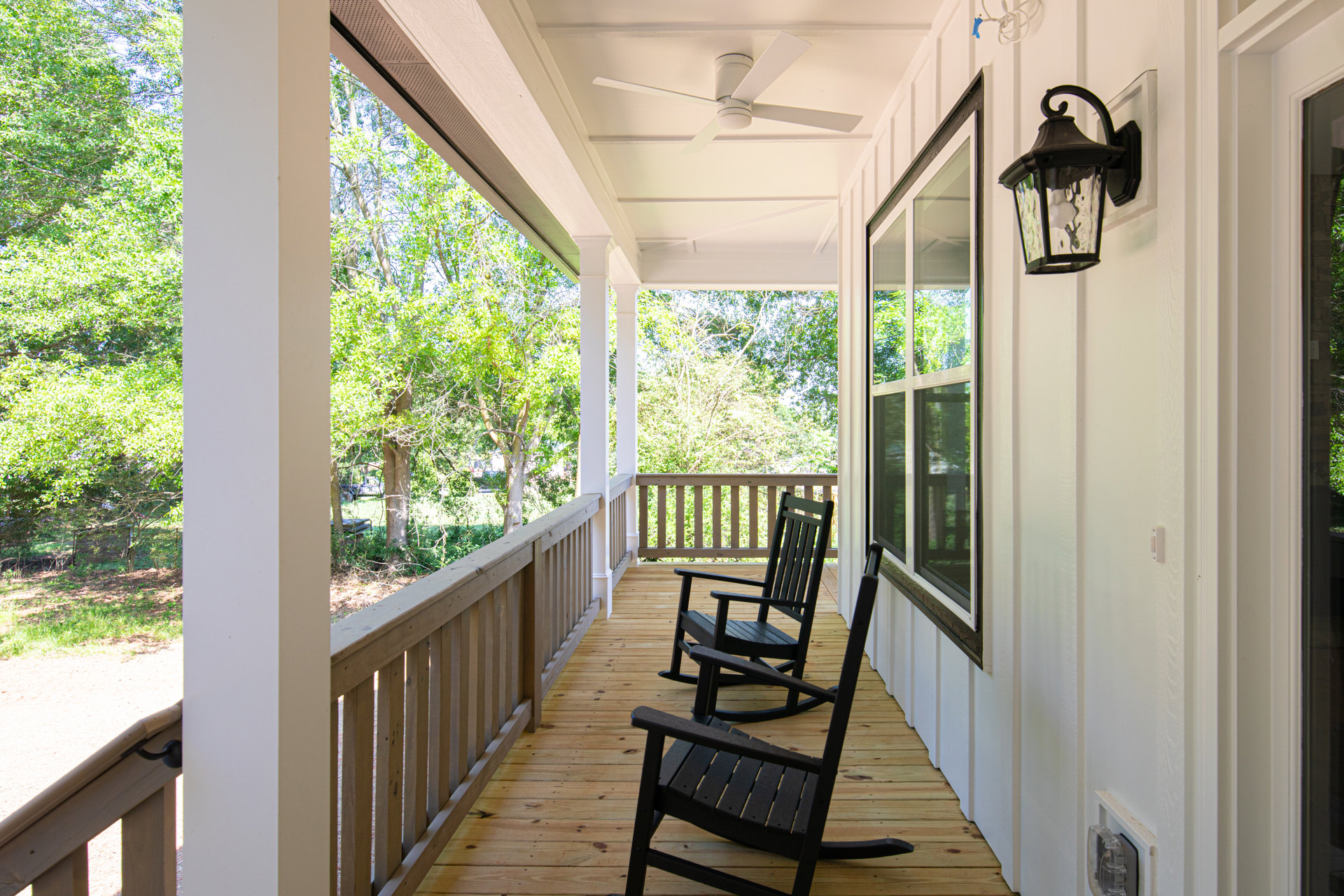 Wooden porch with black rocking chairs, ceiling fan, white railing, and leafy tree in background