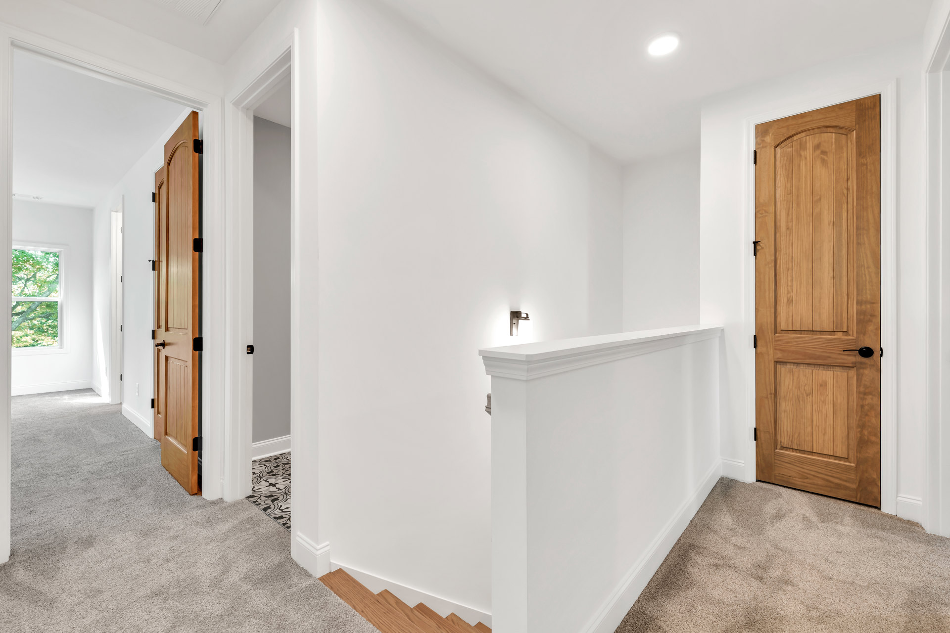 Wood staircase with white risers, adjacent to a wooden door with black handle, white ceiling light, window revealing green trees outside, white walls with wall-mounted light