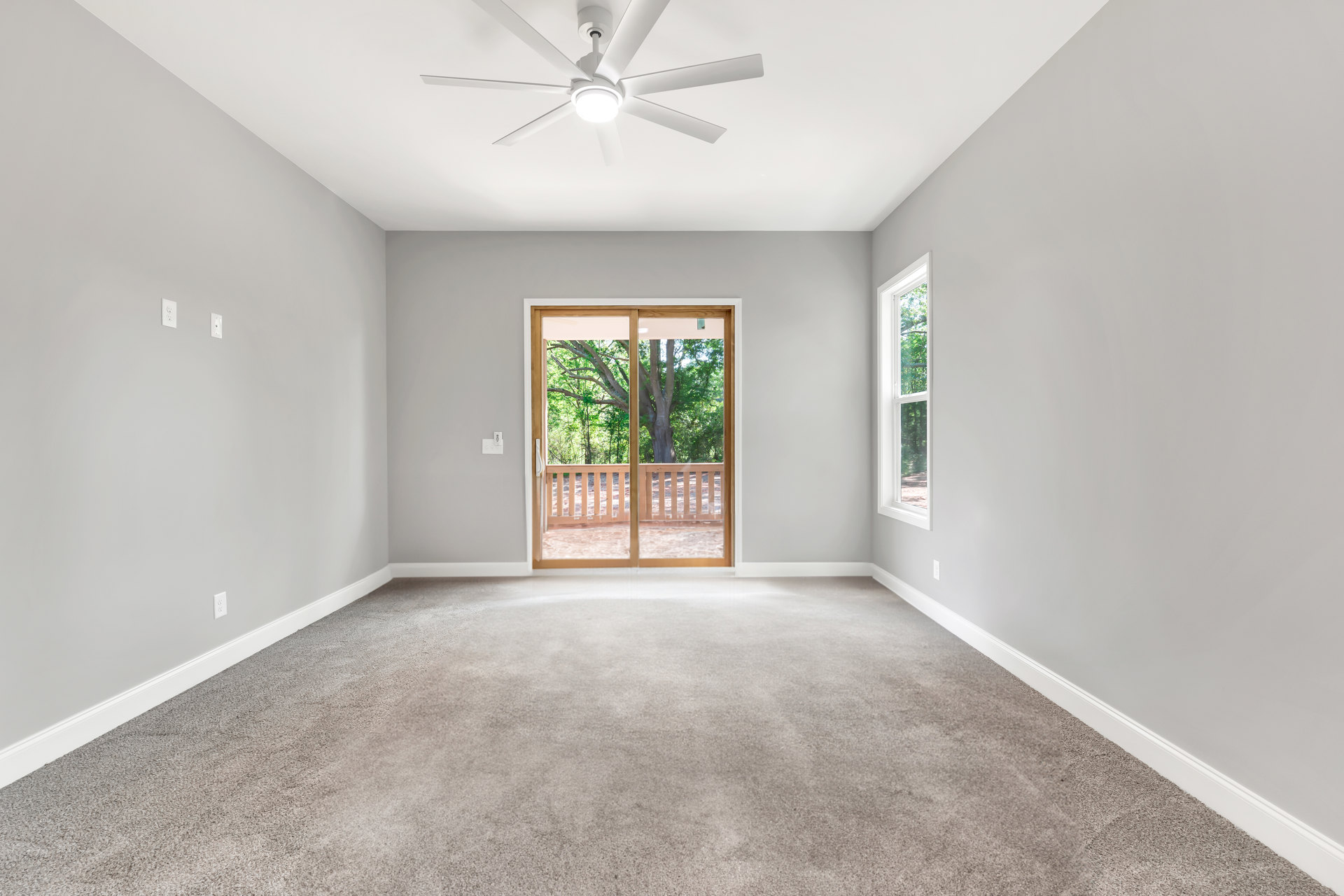 Carpeted room with white ceiling fan and light, large window, sliding glass door overlooking trees, and wooden railing outside