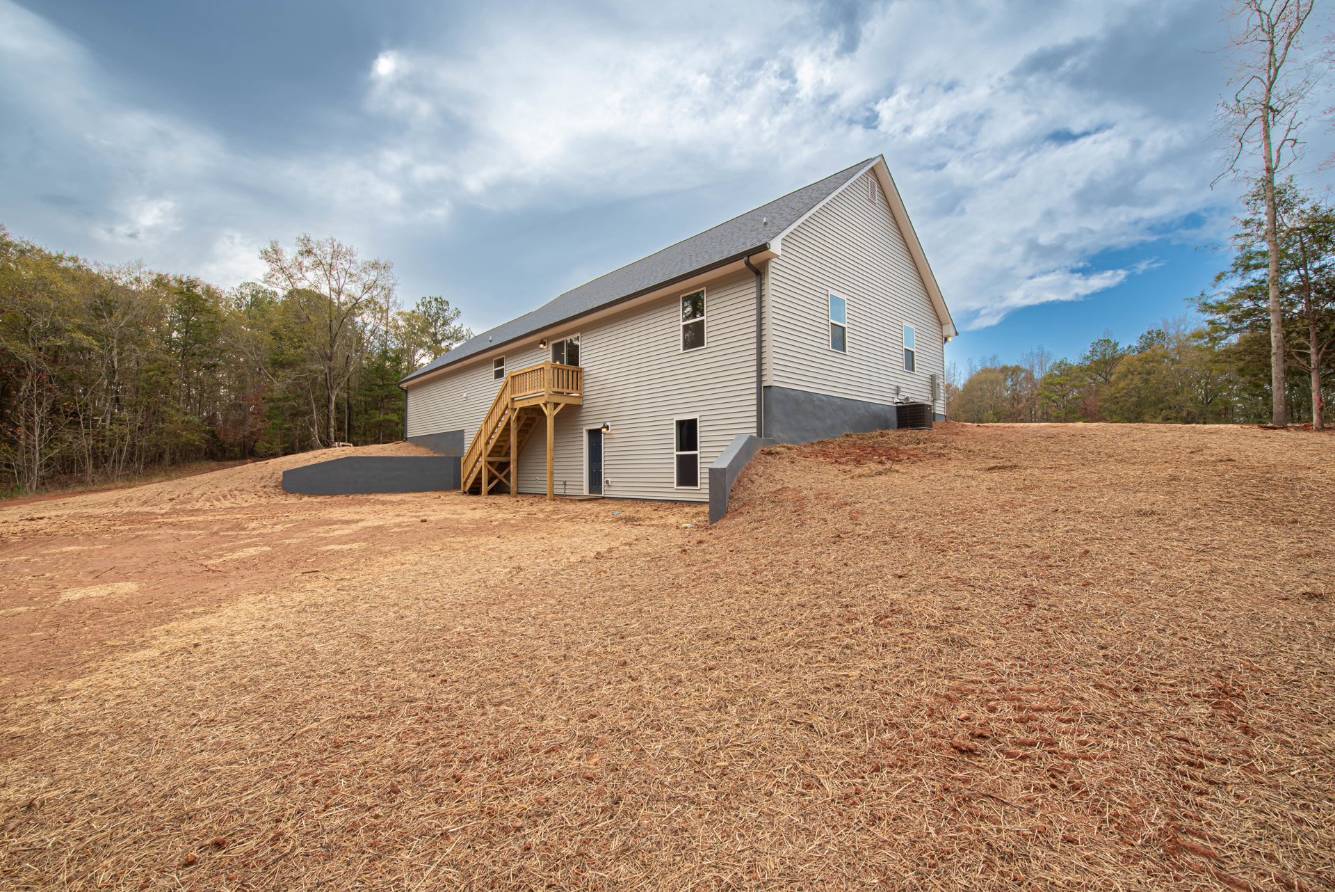 Modern home with sloped roof and elevated wooden deck, exterior staircase leading down to a hill covered in wood chips, surrounded by leafless trees and dry grass under a cloudy