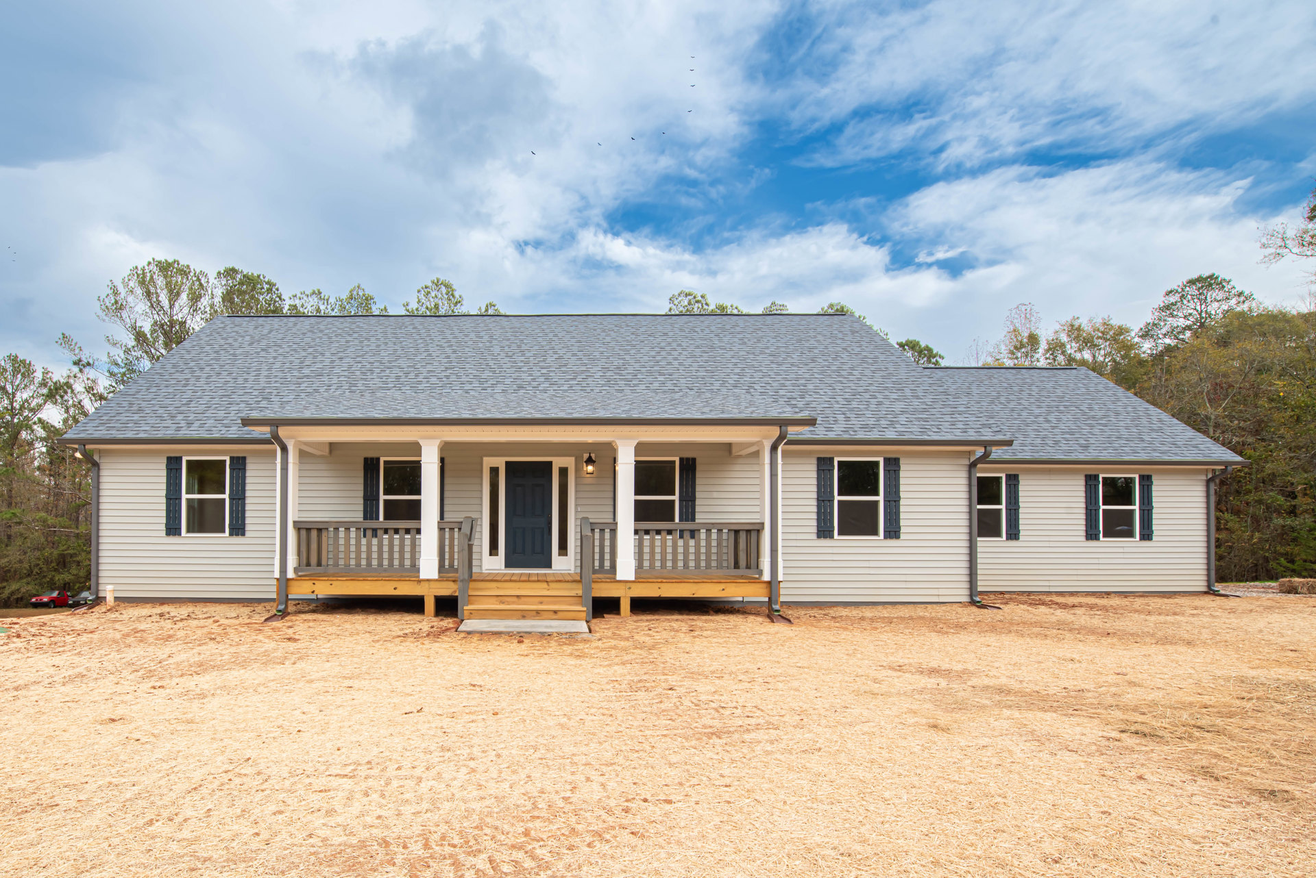 White siding house with wooden porch, blue front door, wide windows, landscaped yard with green grass, leafy tree, and cloudy sky overhead