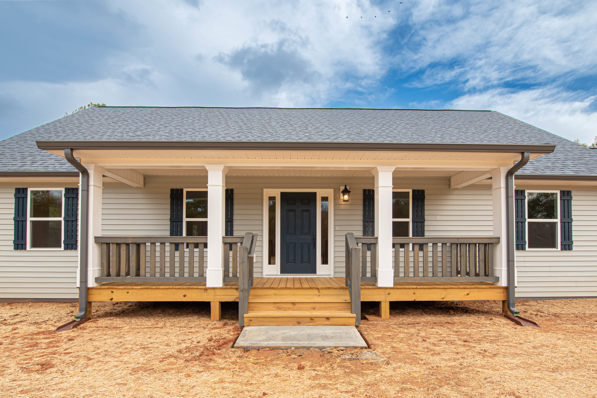 Front porch with wooden steps, blue door, black framed windows, concrete slab, light siding, gabled roof, tree reflection in window, birds in cloudy sky