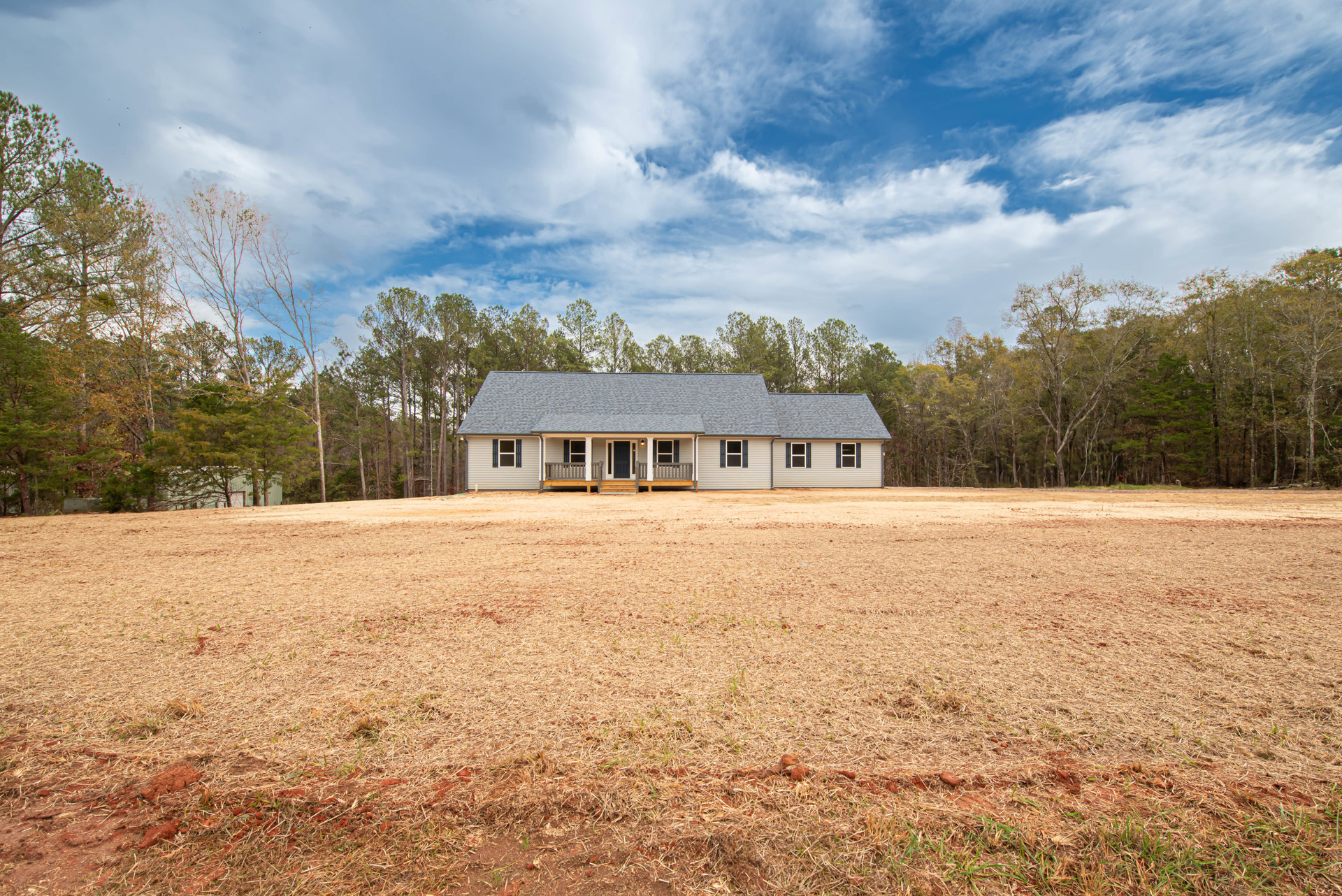 Two-story house with covered porch, surrounded by a wide field of dry brown grass, mature trees, and blue sky with scattered clouds