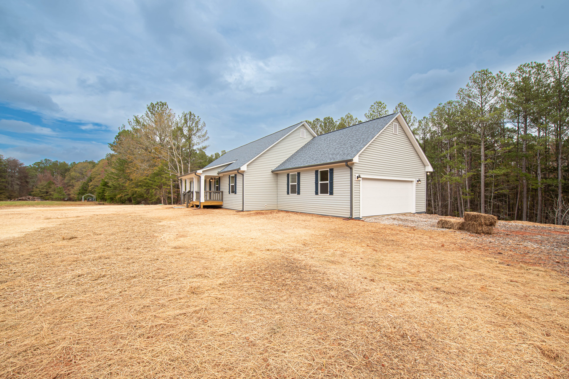 Two-story house with light siding, large grassy lawn, wooden deck, and mature trees in the background under a partly cloudy sky