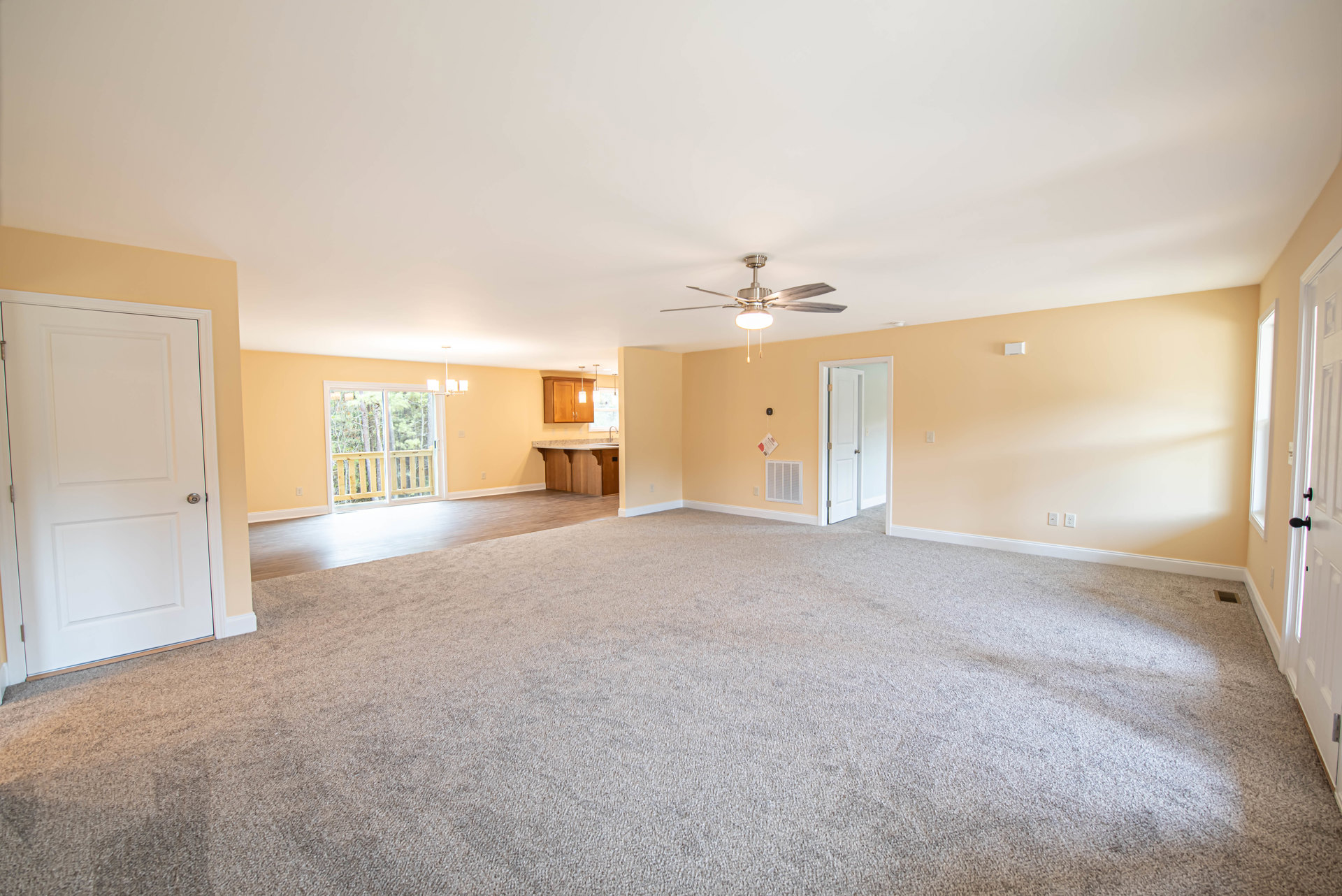 Spacious living room with light carpet flooring, ceiling fan with light fixture, white door featuring silver knob, window overlooking trees with wooden railing, wall vent, and