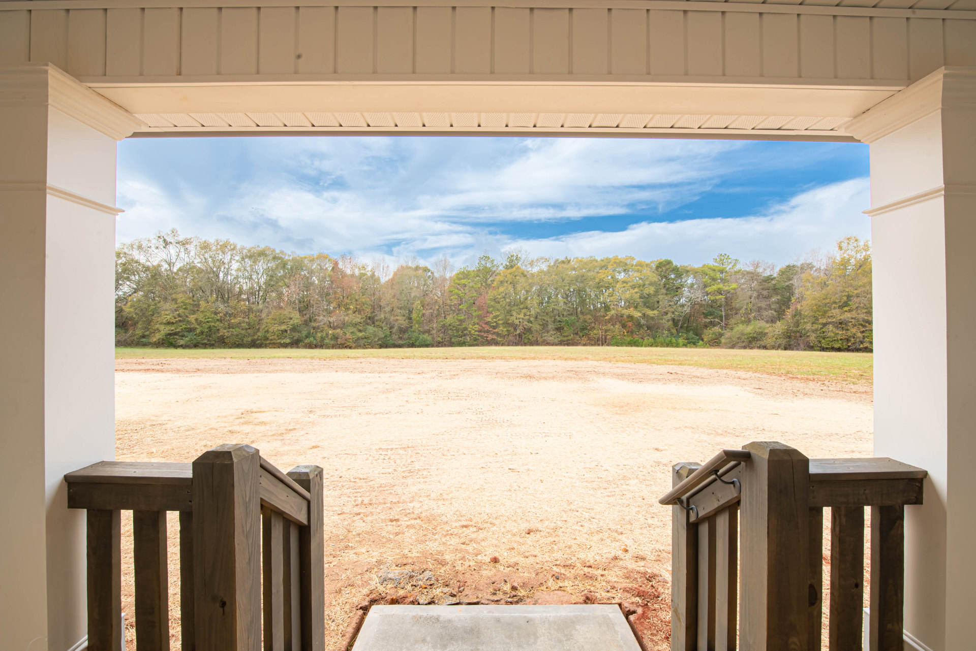 Open garage door framing a dirt field with scattered rocks and grass, wooden porch railing on the left, distant trees and cloudy sky in the background
