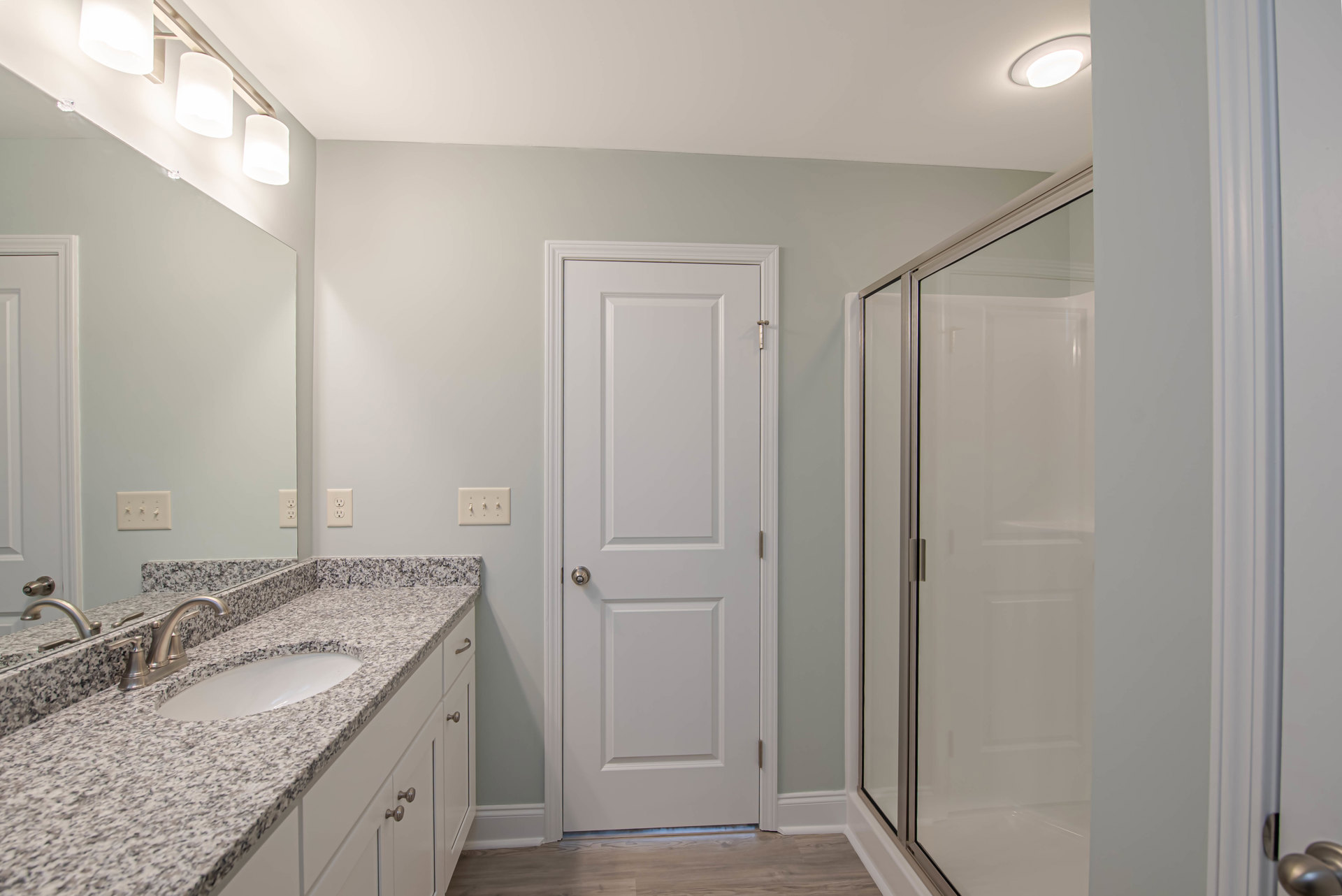 Bathroom with tiled shower, white sink and countertop, wall-mounted mirror, white door with silver knob, three-switch light panel, and white lamp shade.