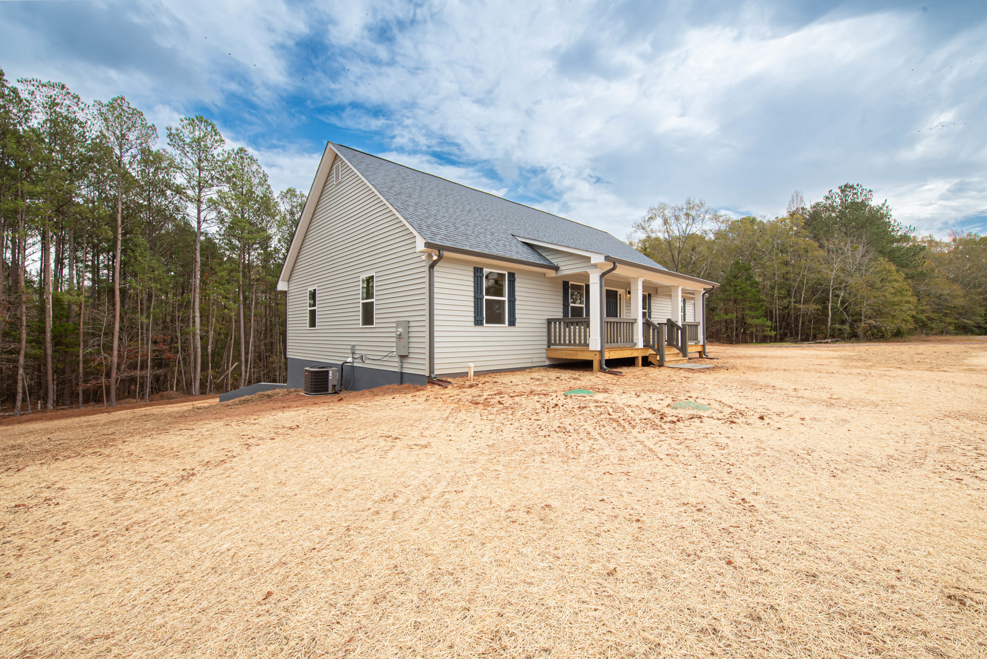 Two-story house with white-framed windows, covered porch, wooden deck, heat pump unit beside exterior wall, dirt yard in foreground, cloudy sky overhead, trees lining property edge