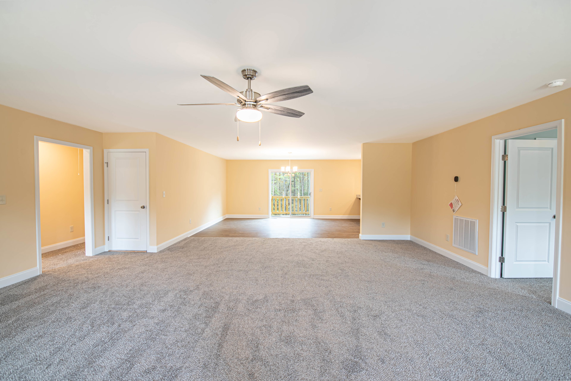 Carpeted bedroom with ceiling fan and light, white walls, two doors with brass and black handles, window overlooking trees