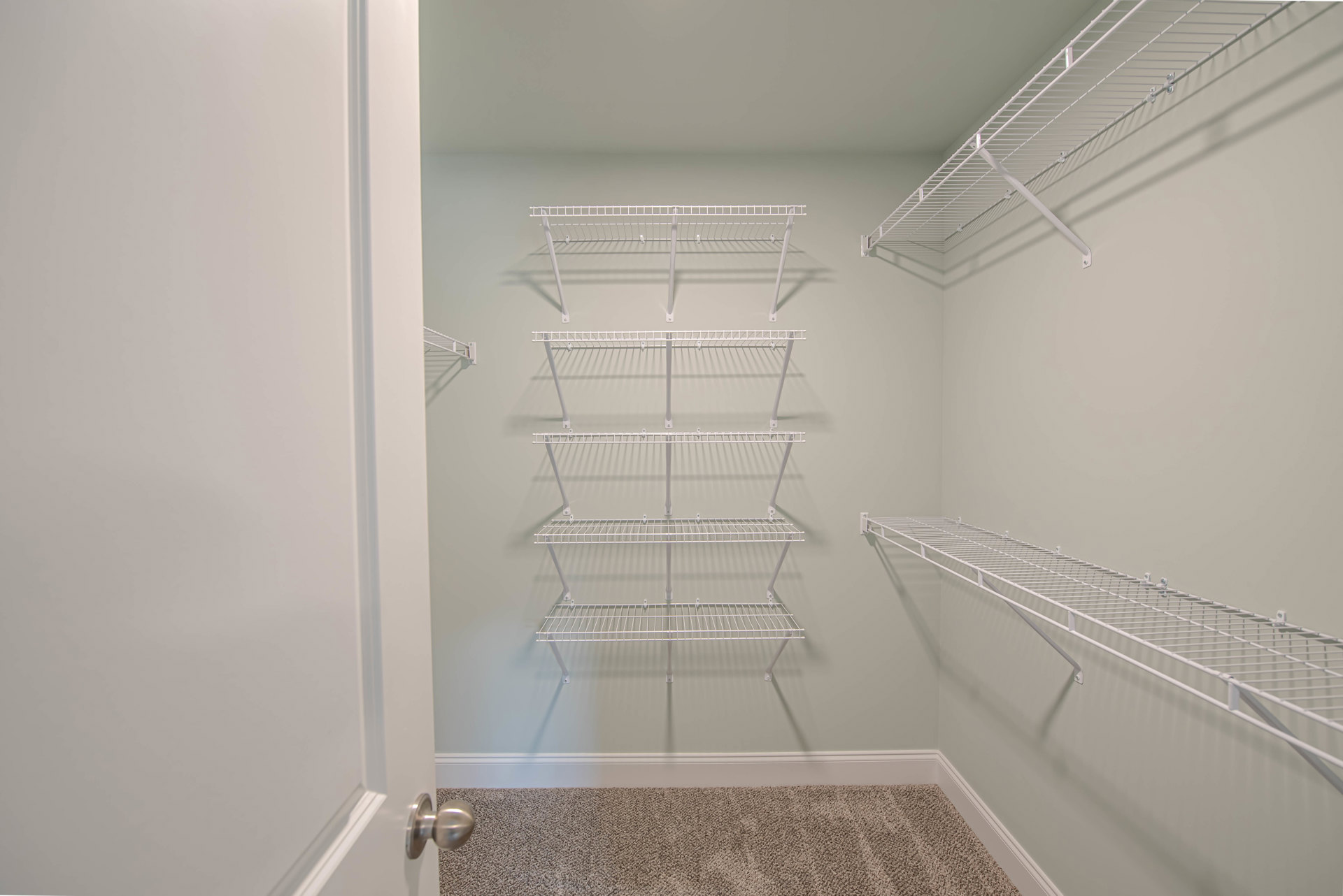 Walk-in closet with white wire shelves mounted on plaster walls, beige carpet flooring, and a close-up view of a brushed metal door knob.