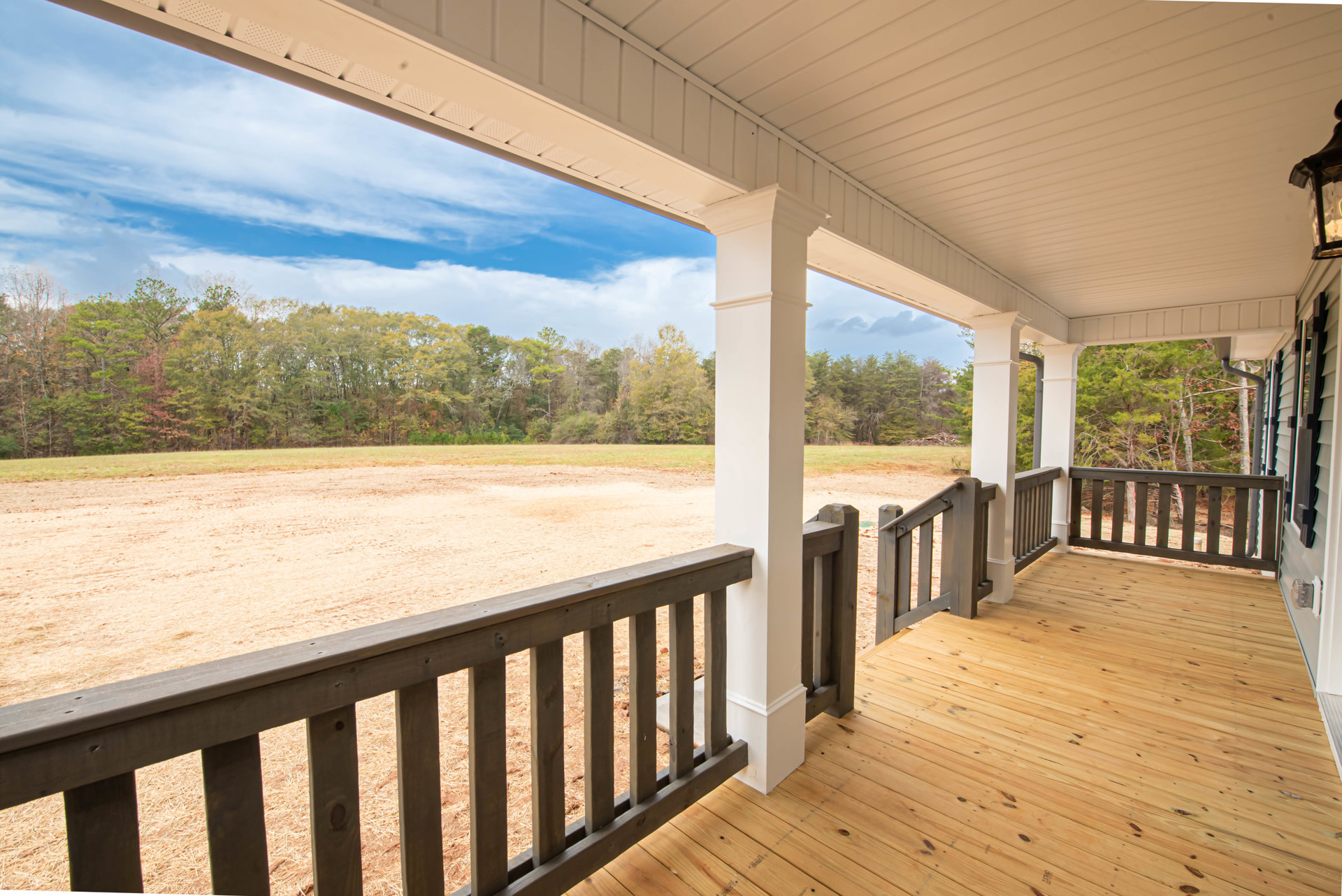 White porch with wooden railing overlooking open field and scattered trees under blue sky with clouds