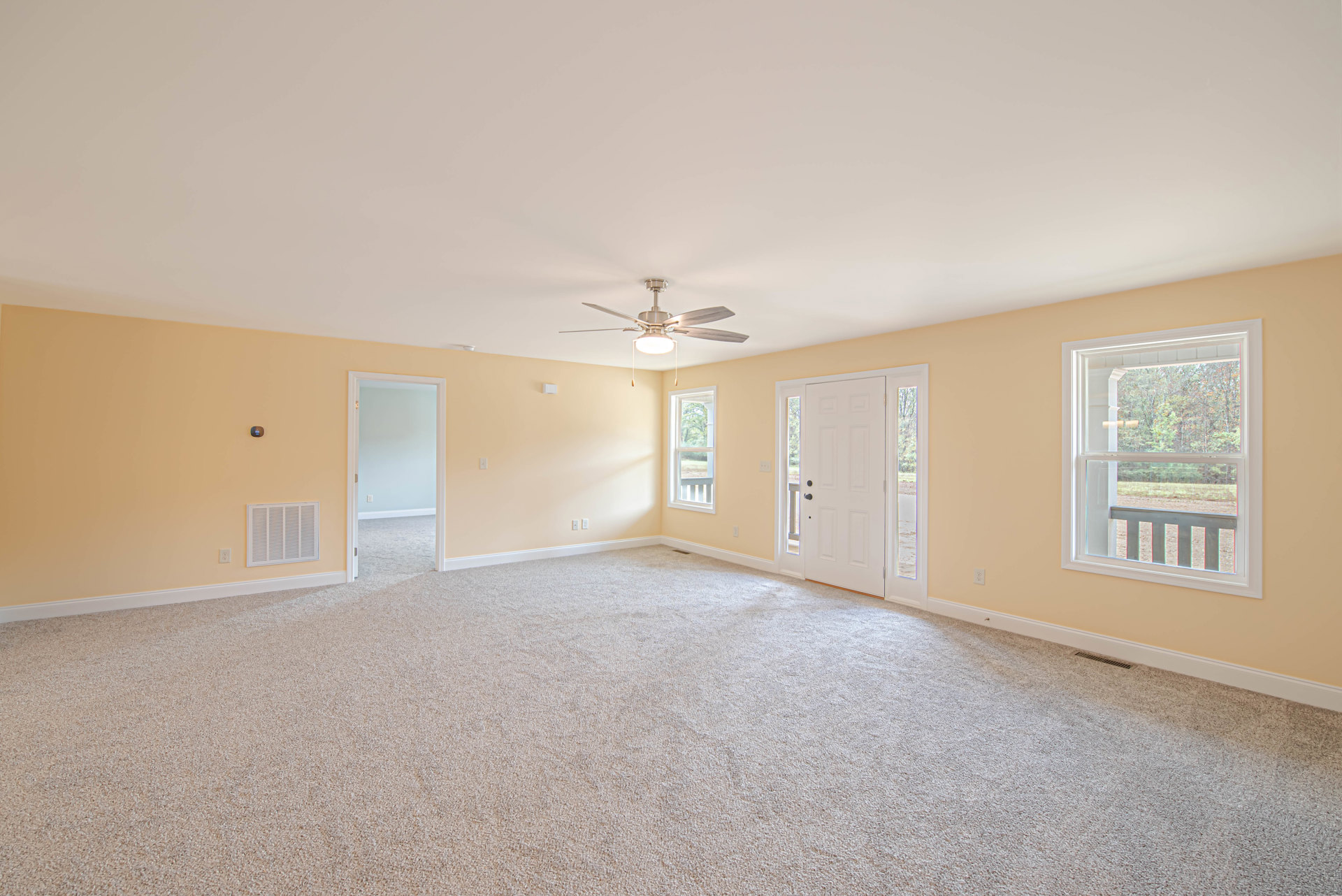 Carpeted room with white walls, ceiling fan with light, large window showing trees outside, white door with black knobs, white door frame, simple molding along ceiling and floor.