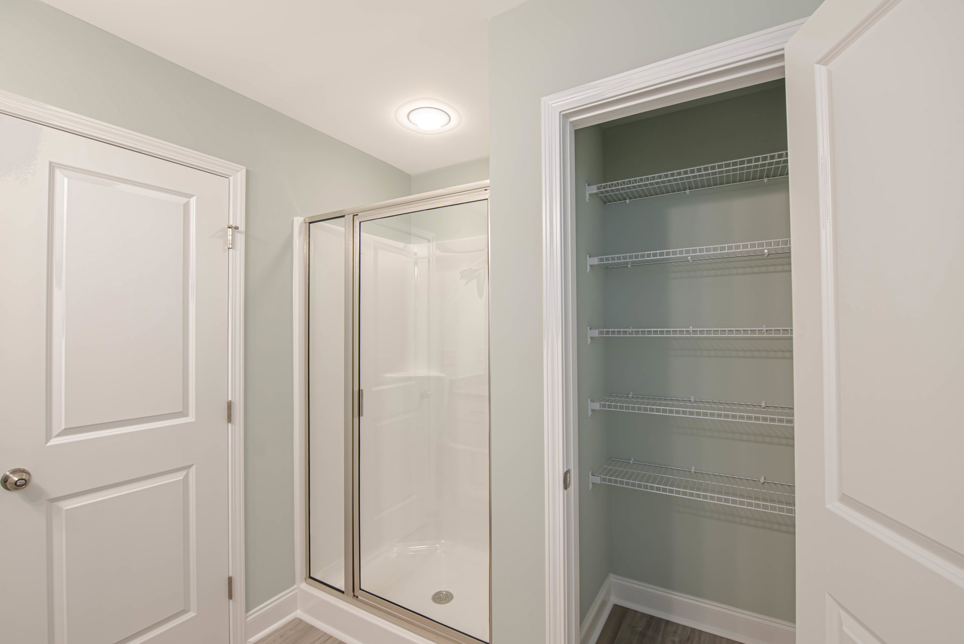 Bathroom with glass shower door, built-in white shelving with metal brackets, white paneled door with chrome handle, recessed ceiling light, plaster walls, and crown molding.