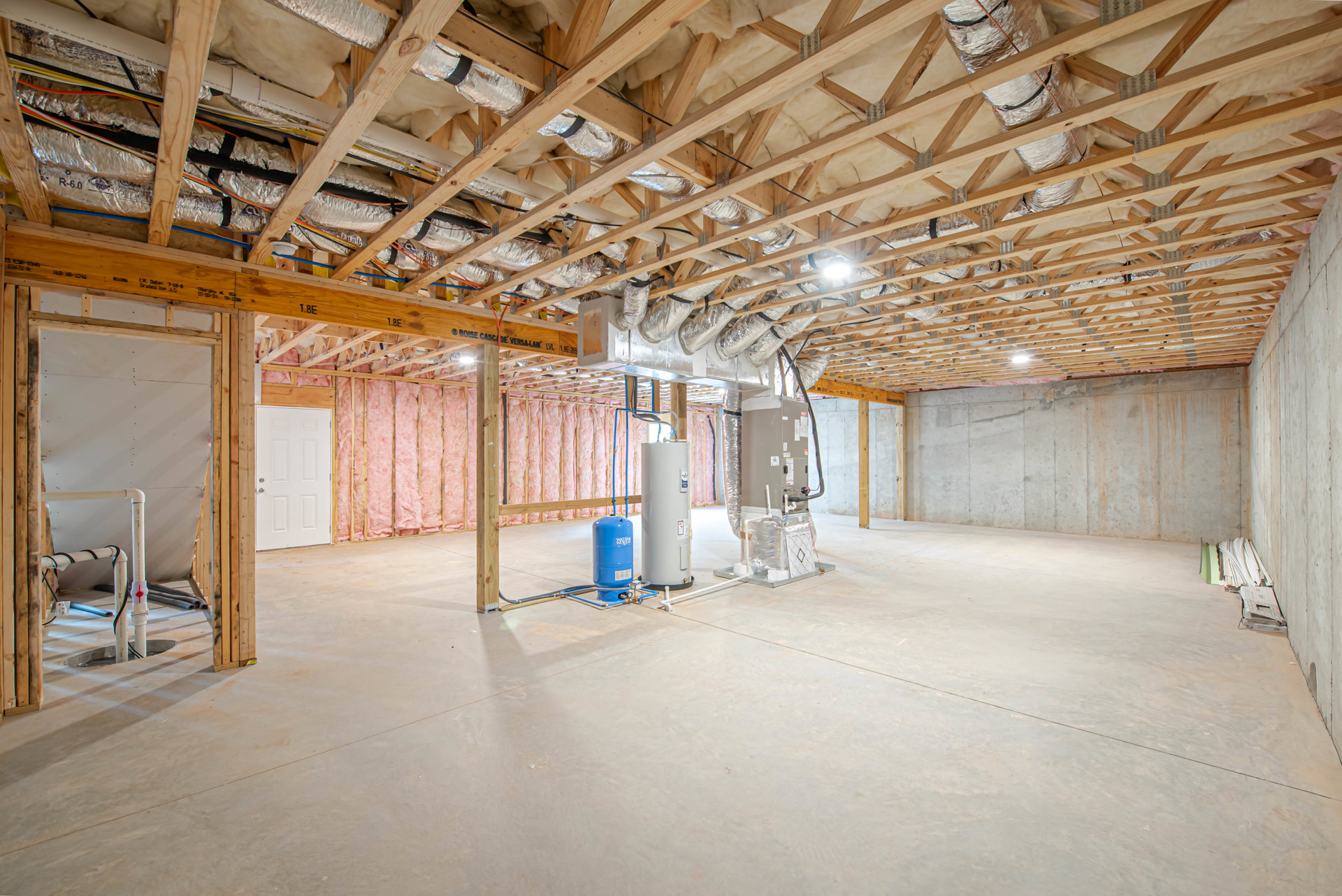 Utility room with exposed wooden ceiling beams, metal pipes, large white door with silver handle, blue barrel with white text, and concrete floor