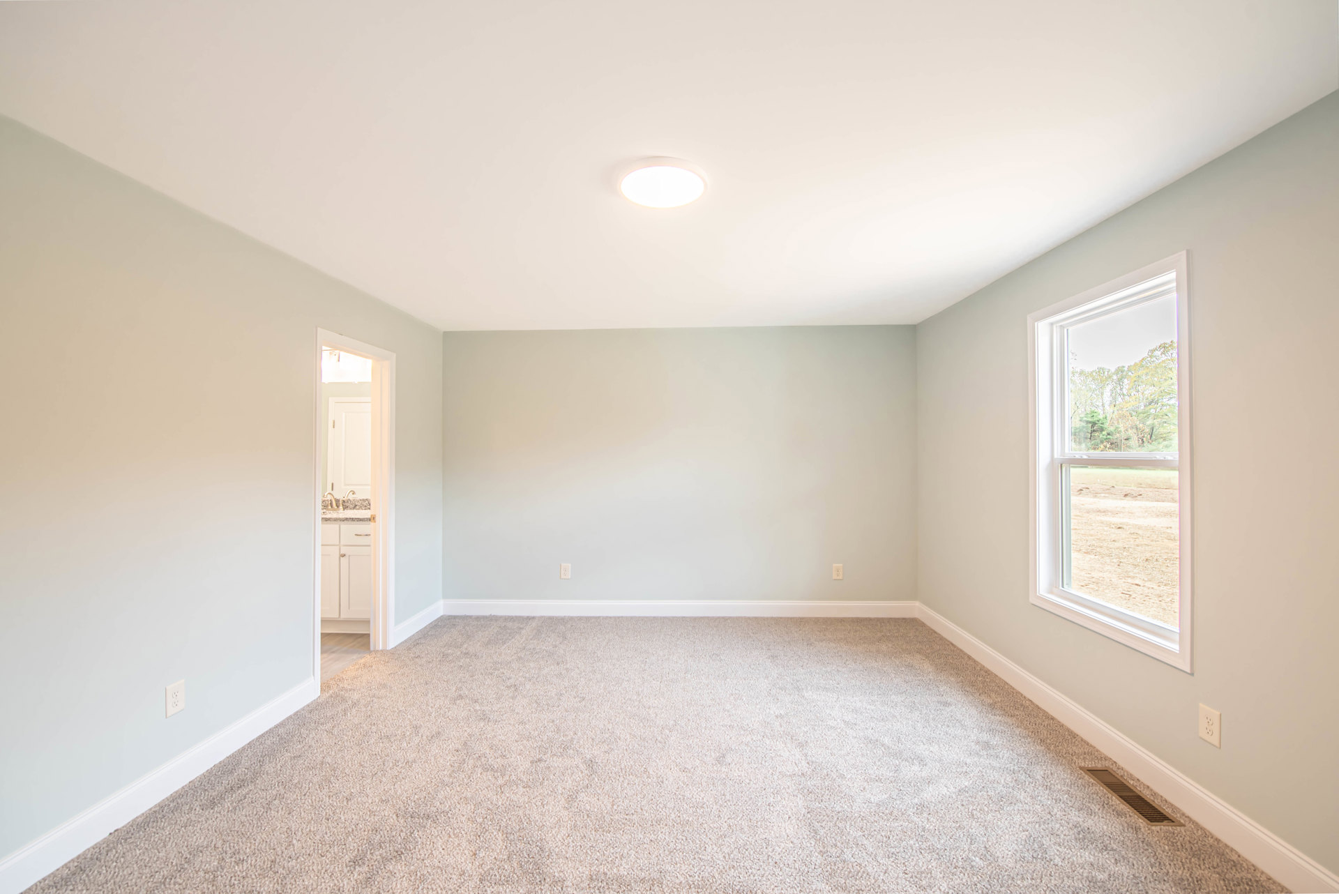 Neutral-toned carpeted bedroom with white walls, recessed ceiling light, rectangular window overlooking leafy trees, floor vent near baseboard molding