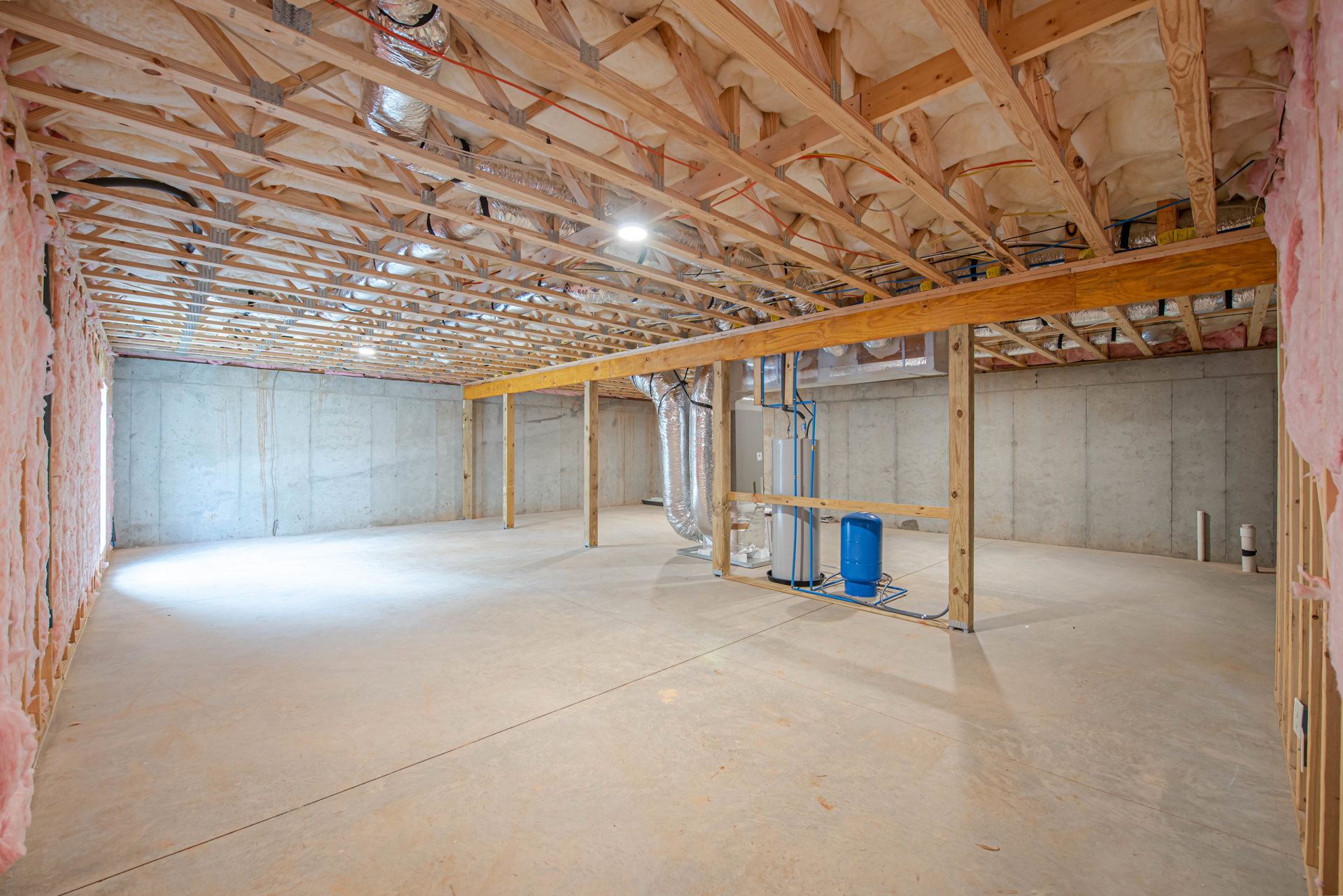 Concrete floor and ceiling with exposed wooden beams, metal pipes, blue barrel on metal stand, white ceiling material, and overhead light fixture.