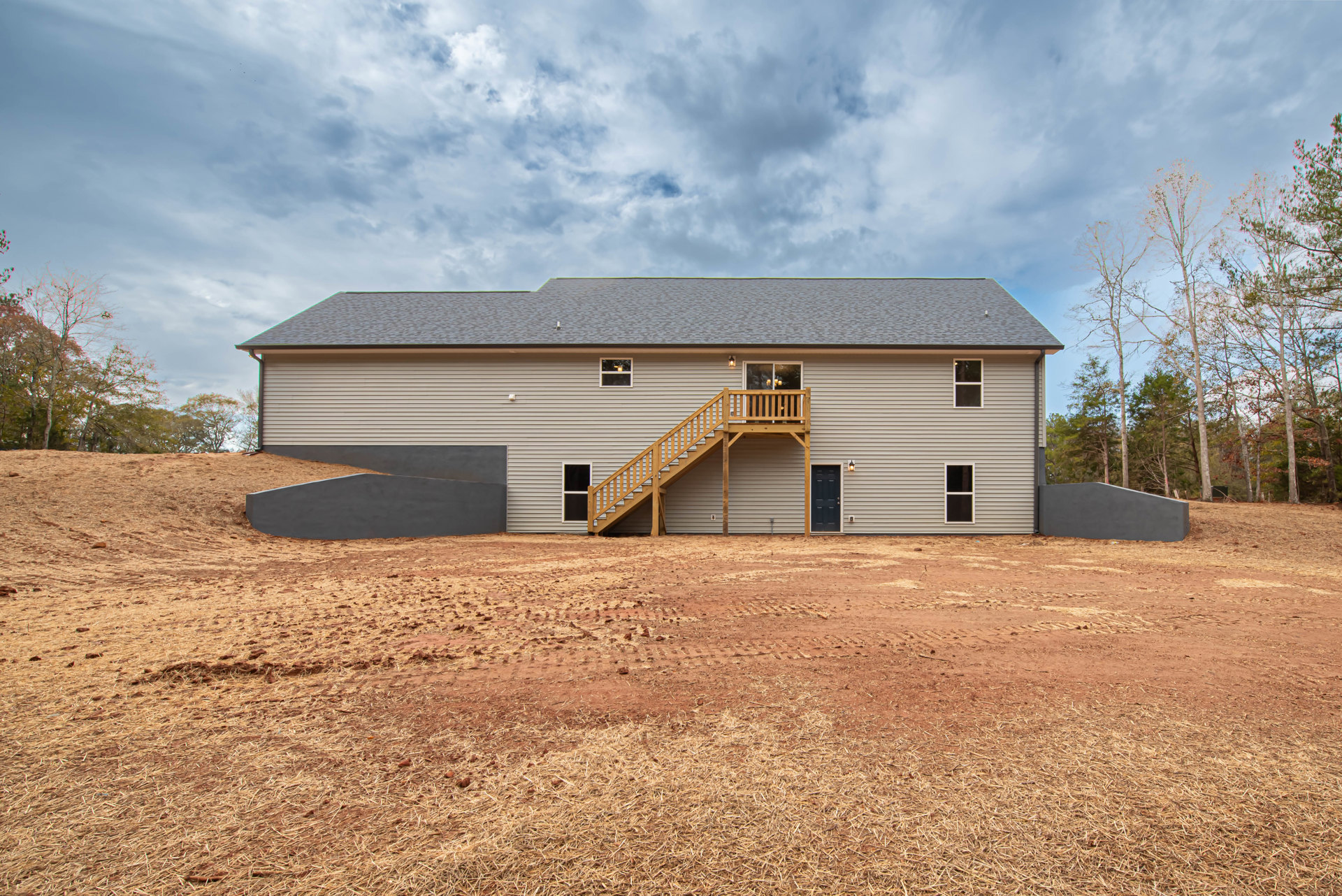 Two-story home with wood deck and staircase leading down to dirt yard, gray siding, gabled roof, and cloudy sky overhead