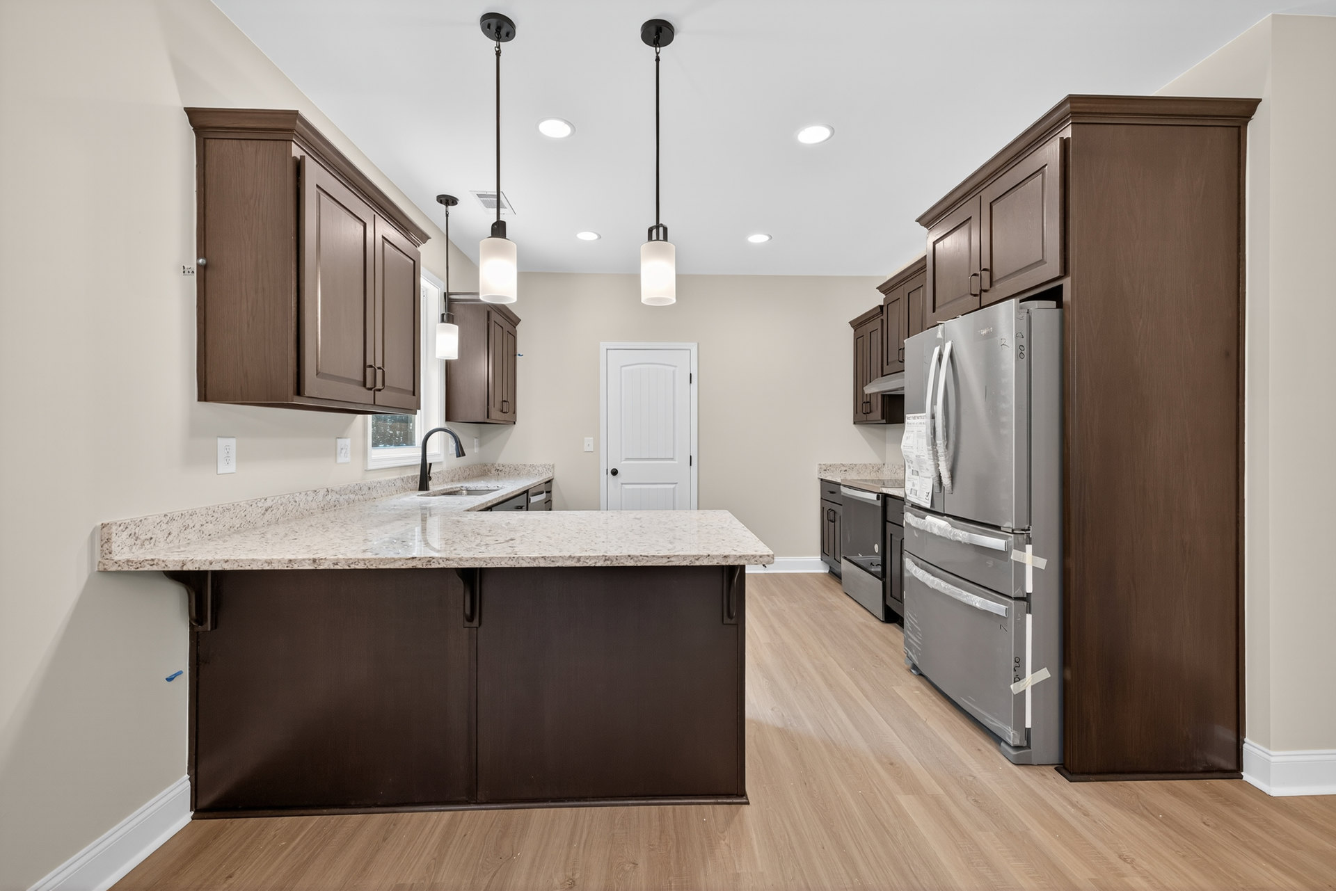 Spacious kitchen featuring a large central island with stone countertop, stainless steel refrigerator partially wrapped in plastic, white cabinetry with black knobs, brown cabinet