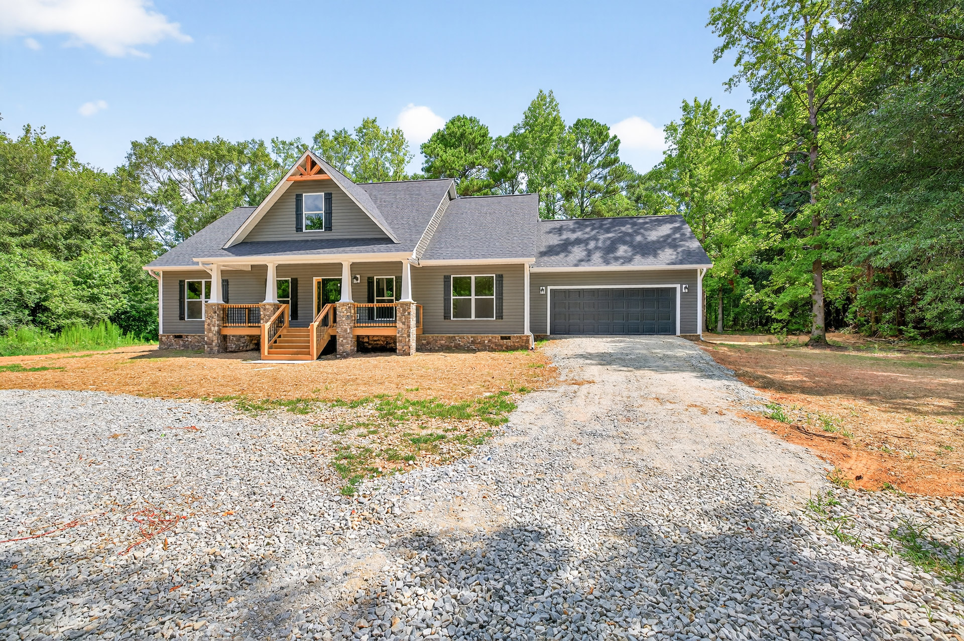 Two-story home with white-framed windows, wooden porch and stairs, stone garage, gravel driveway, and mature trees in the background