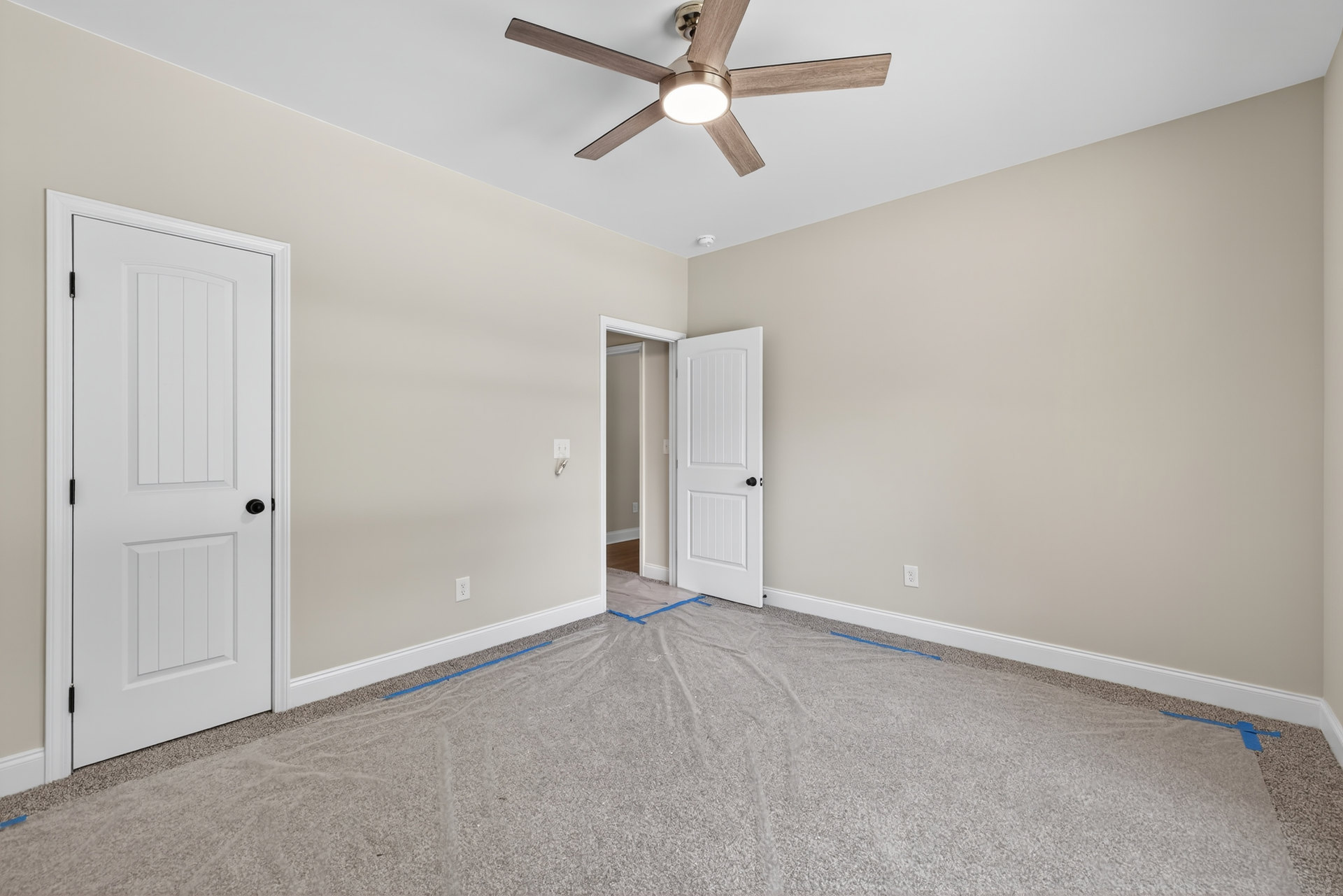 Ceiling fan with integrated light fixture mounted above carpeted floor, white paneled door with black hardware, blue painter’s tape visible along baseboard, neutral walls and