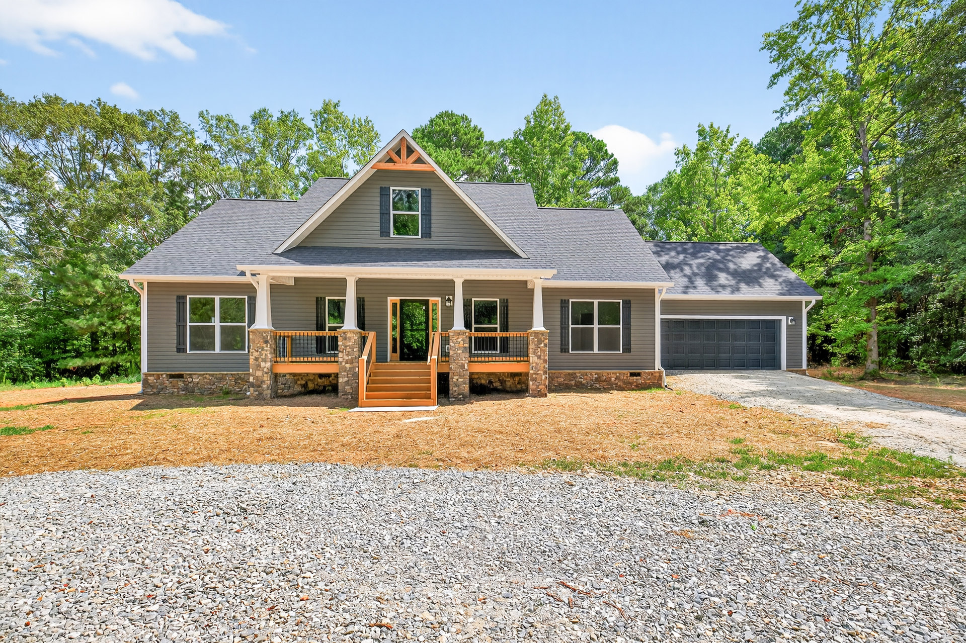Two-story home with white-framed windows, wooden porch and stairs, attached garage with paneled door, concrete driveway, mature trees in the background