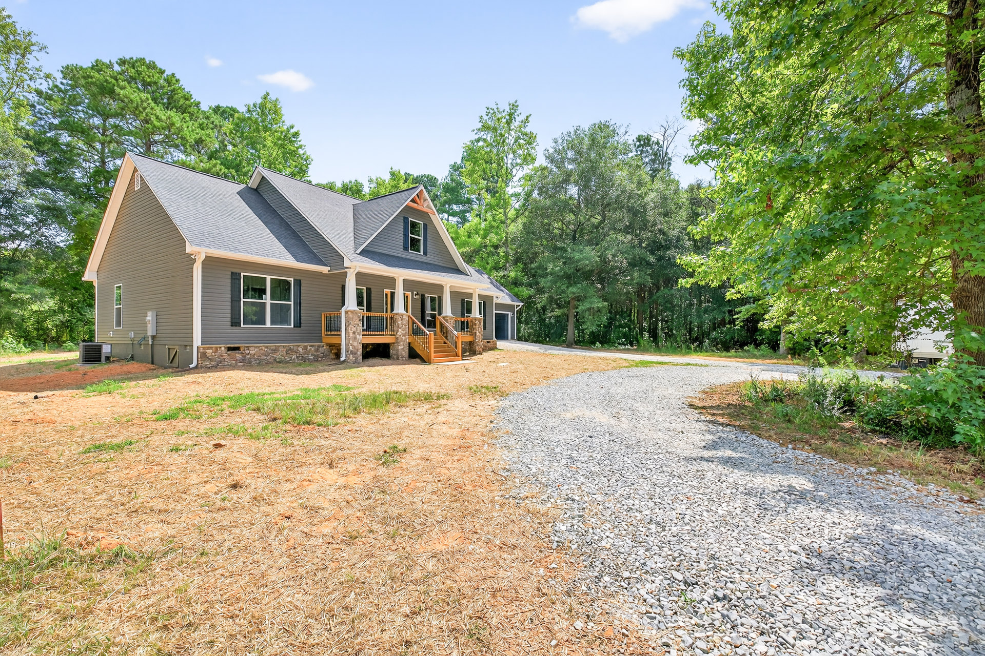 Two-story house with white-framed windows, stone porch wall, gravel driveway, and mature trees with green leaves