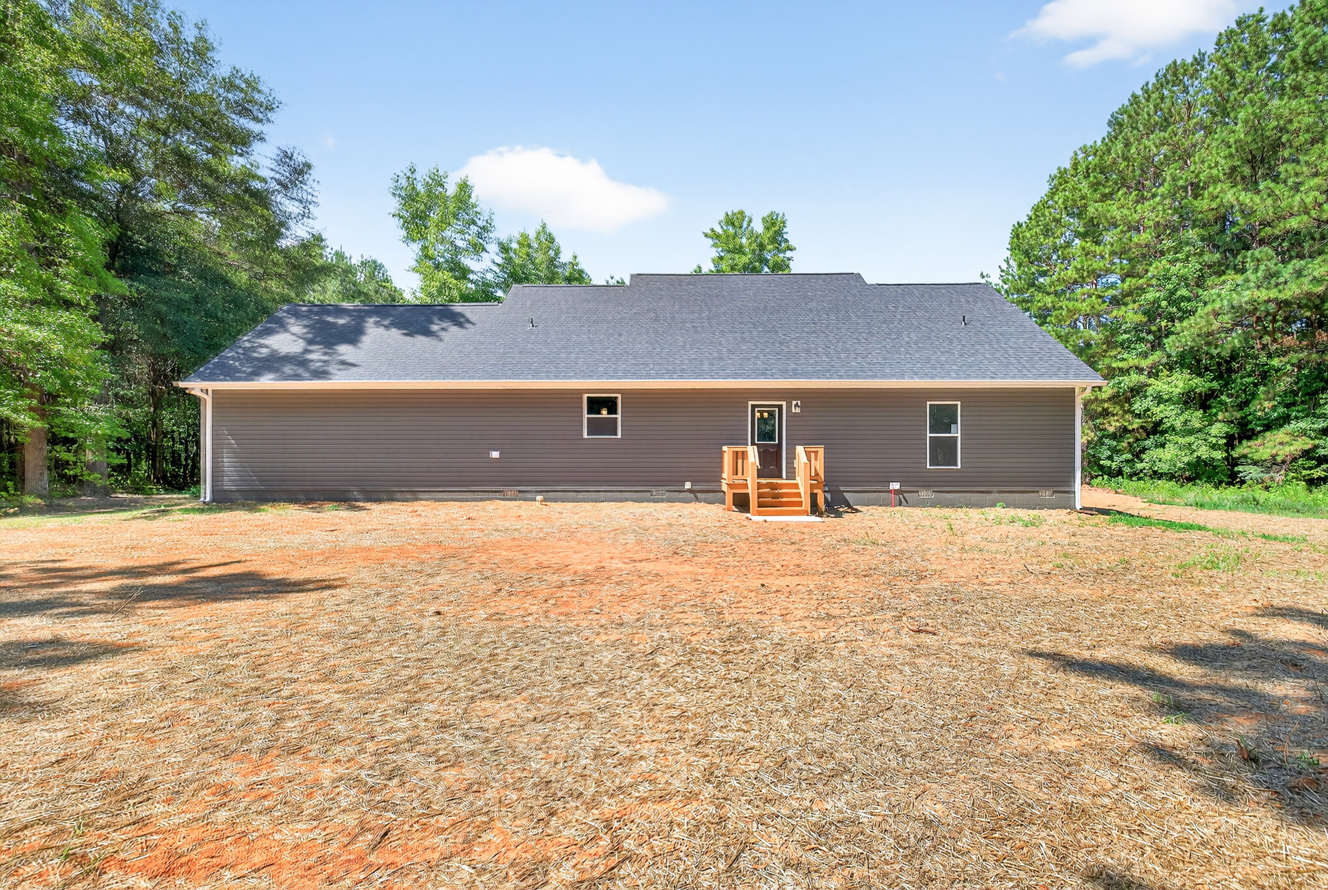Two-story house with gray siding, covered front porch, wooden stairs, landscaped yard, mature trees, and blue sky with scattered clouds