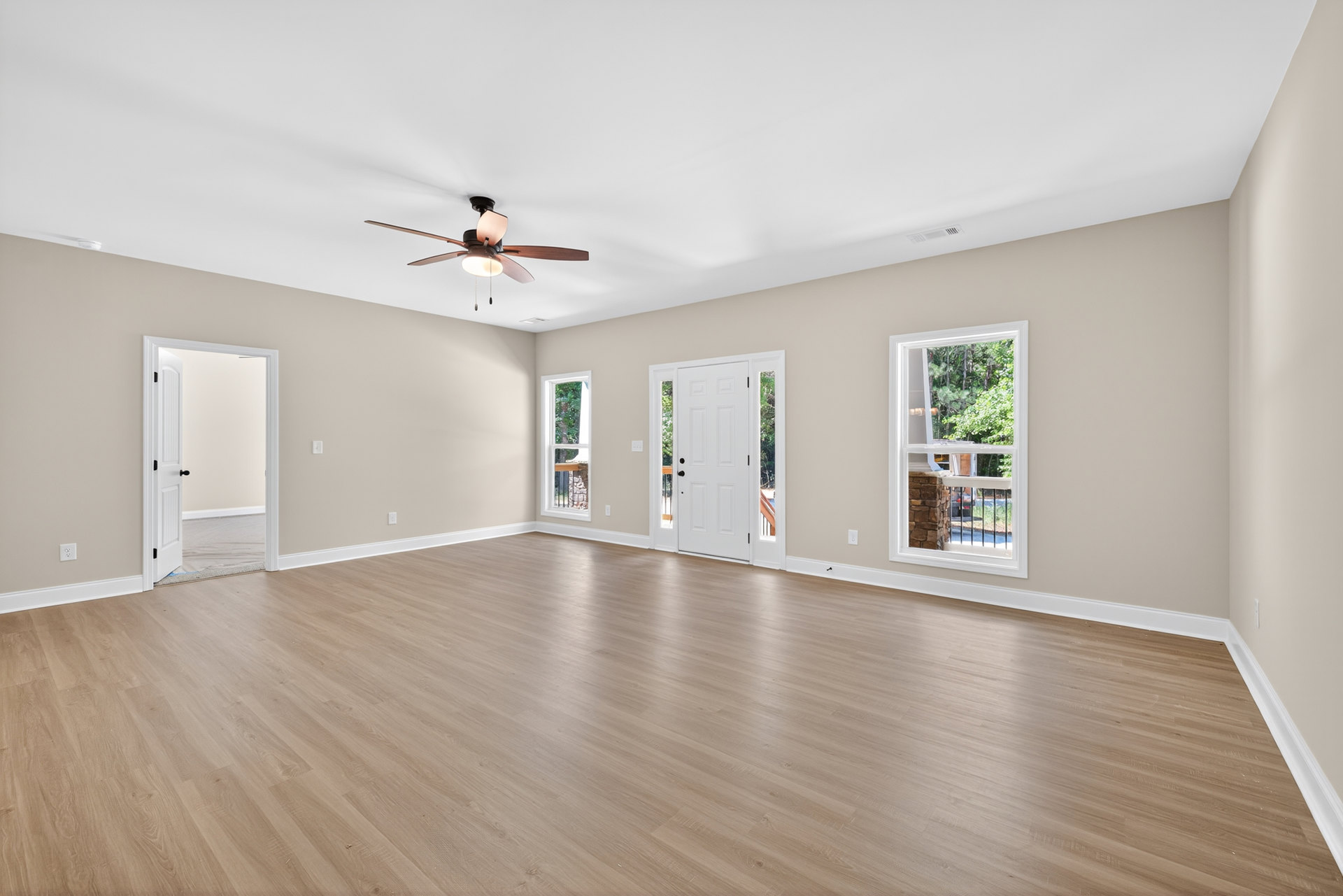 Living room with wood flooring, ceiling fan with light, stone fireplace, large window overlooking trees, and white door with black handle