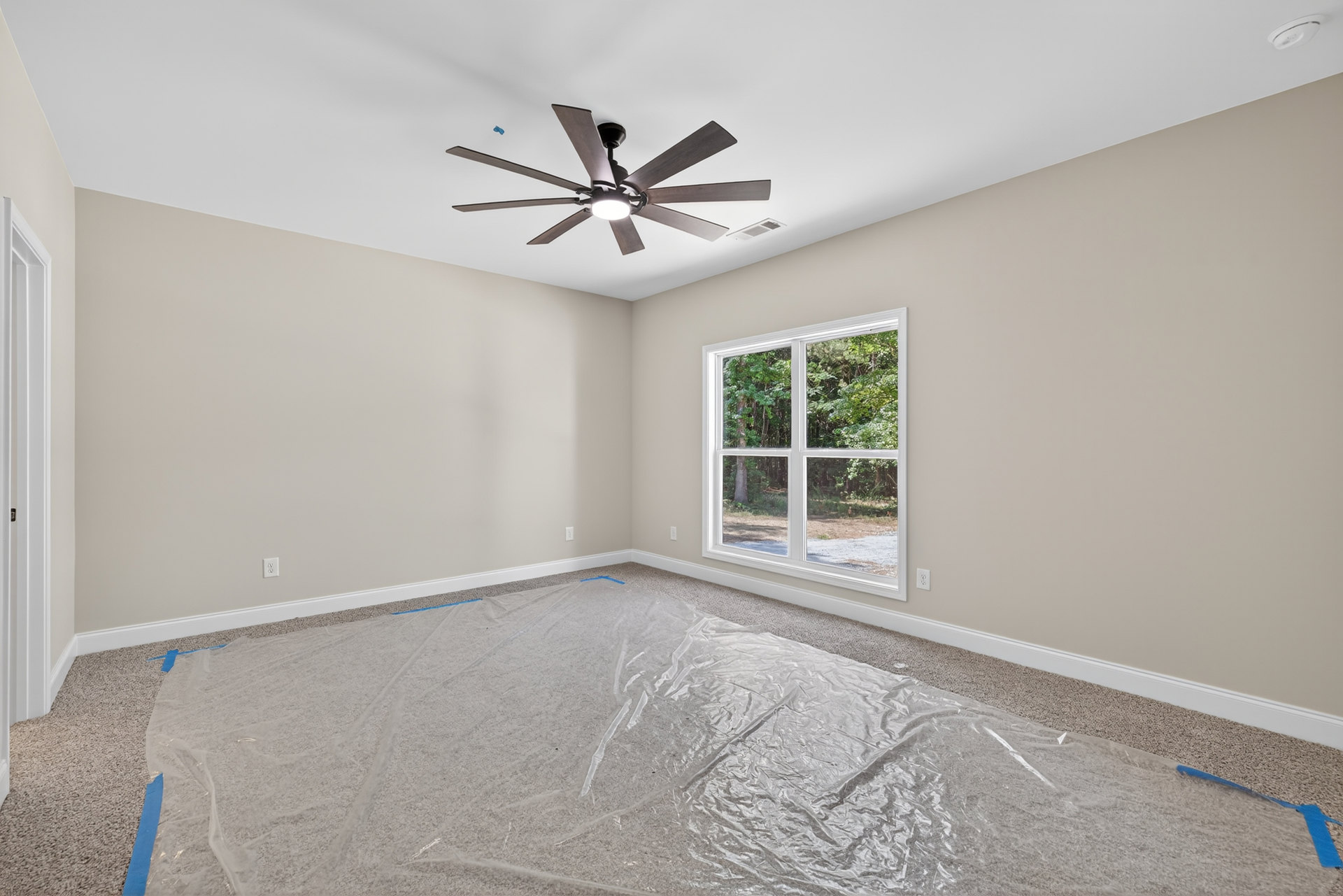 Ceiling fan with integrated light fixture mounted on white plaster ceiling above room with plastic covering the floor, window revealing trees outside, close-up view of door and