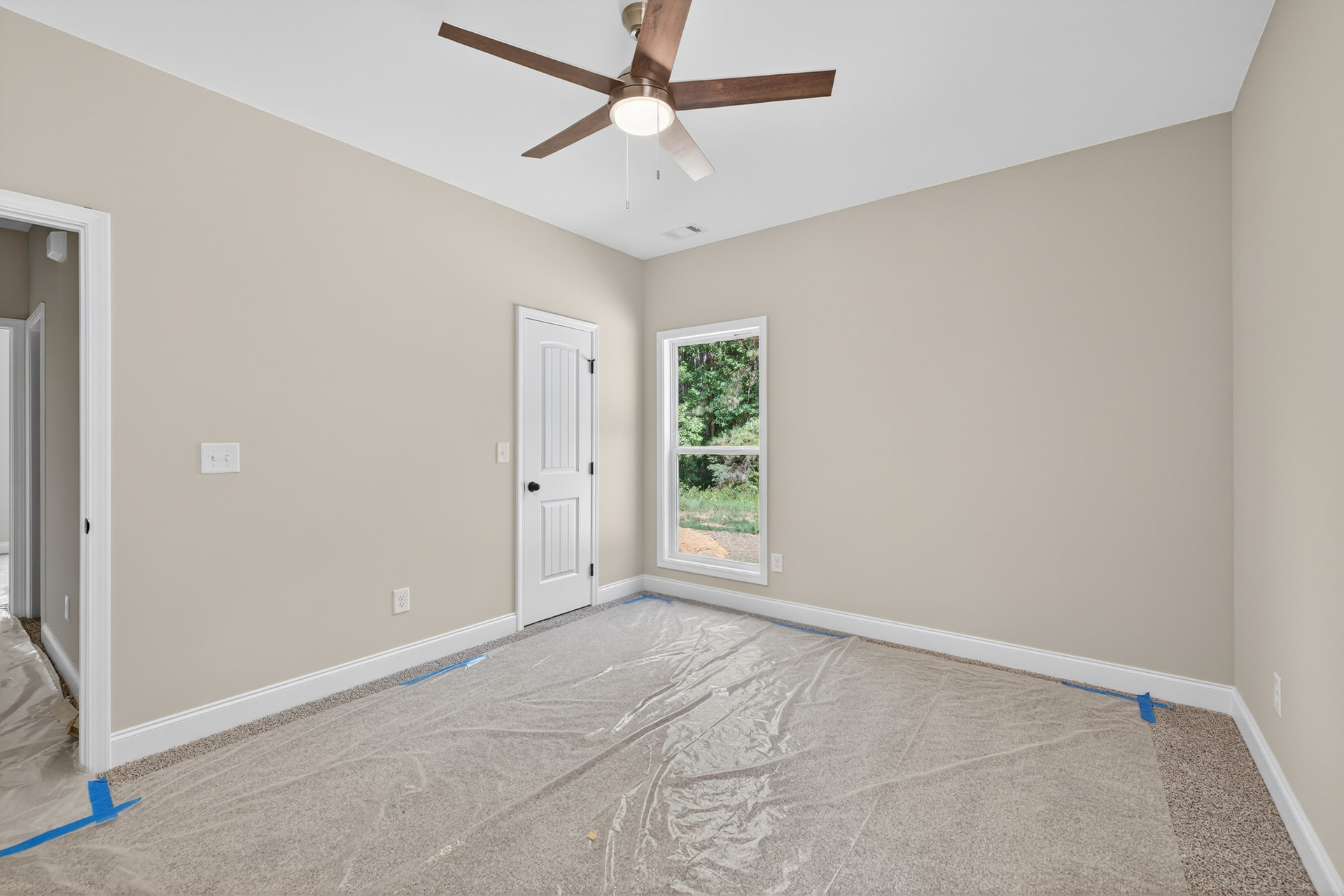 Ceiling fan with light fixture mounted on white ceiling above room with white door featuring black knobs, window showing trees outside, plastic covering on floor, close-up of