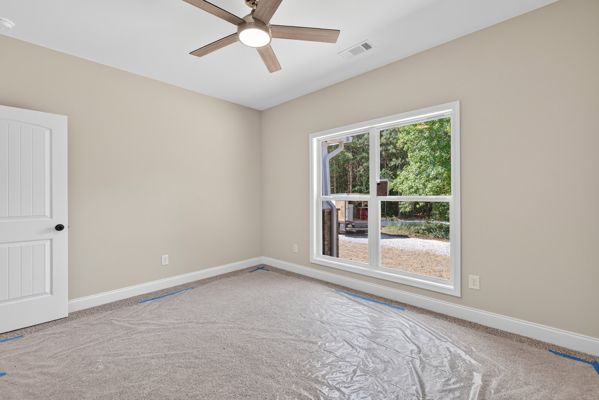 Ceiling fan with light fixture mounted on white plaster ceiling above carpeted floor covered in protective plastic, adjacent to a wooden door and window overlooking a parked truck.