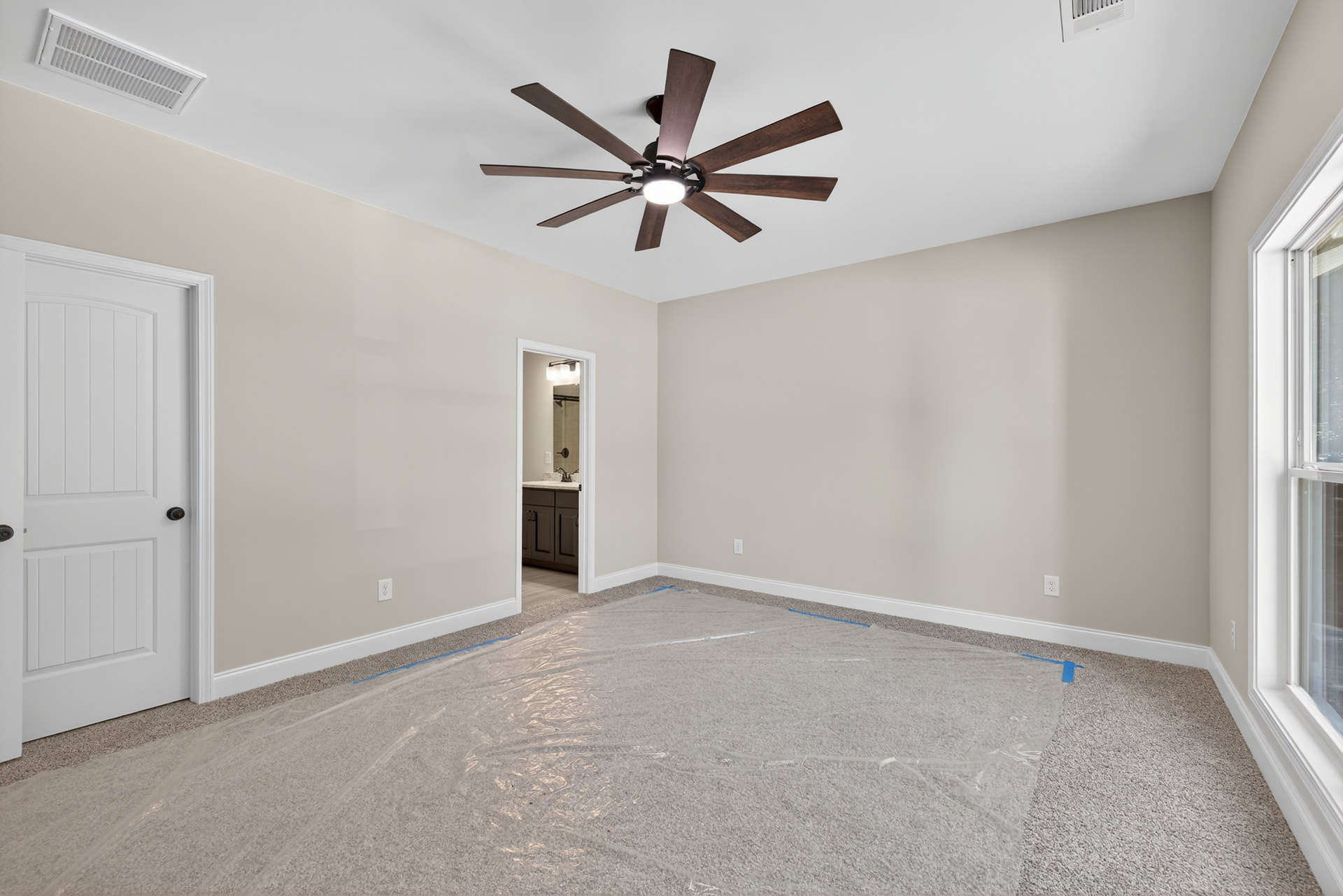 Ceiling fan with light fixture mounted on white ceiling, white door with black knobs, air vent, carpet covered in protective plastic, bathroom sink and mirror visible in adjoining