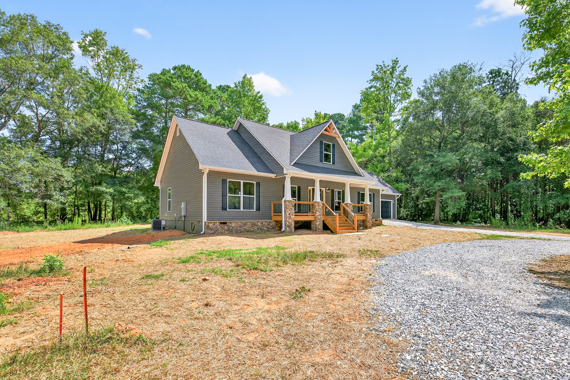 Two-story house with stone wall and white-framed windows, covered porch with railing, gravel driveway bordered by grass, mature trees, and green plants.
