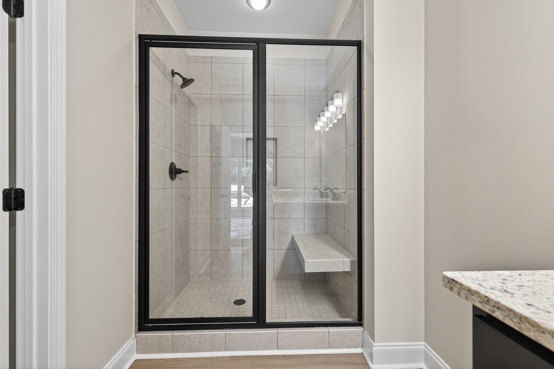 Walk-in shower with glass doors, built-in bench topped with white tile, wall-mounted shower head, black hinge on white tiled surface, partial view of bathroom countertop and sink