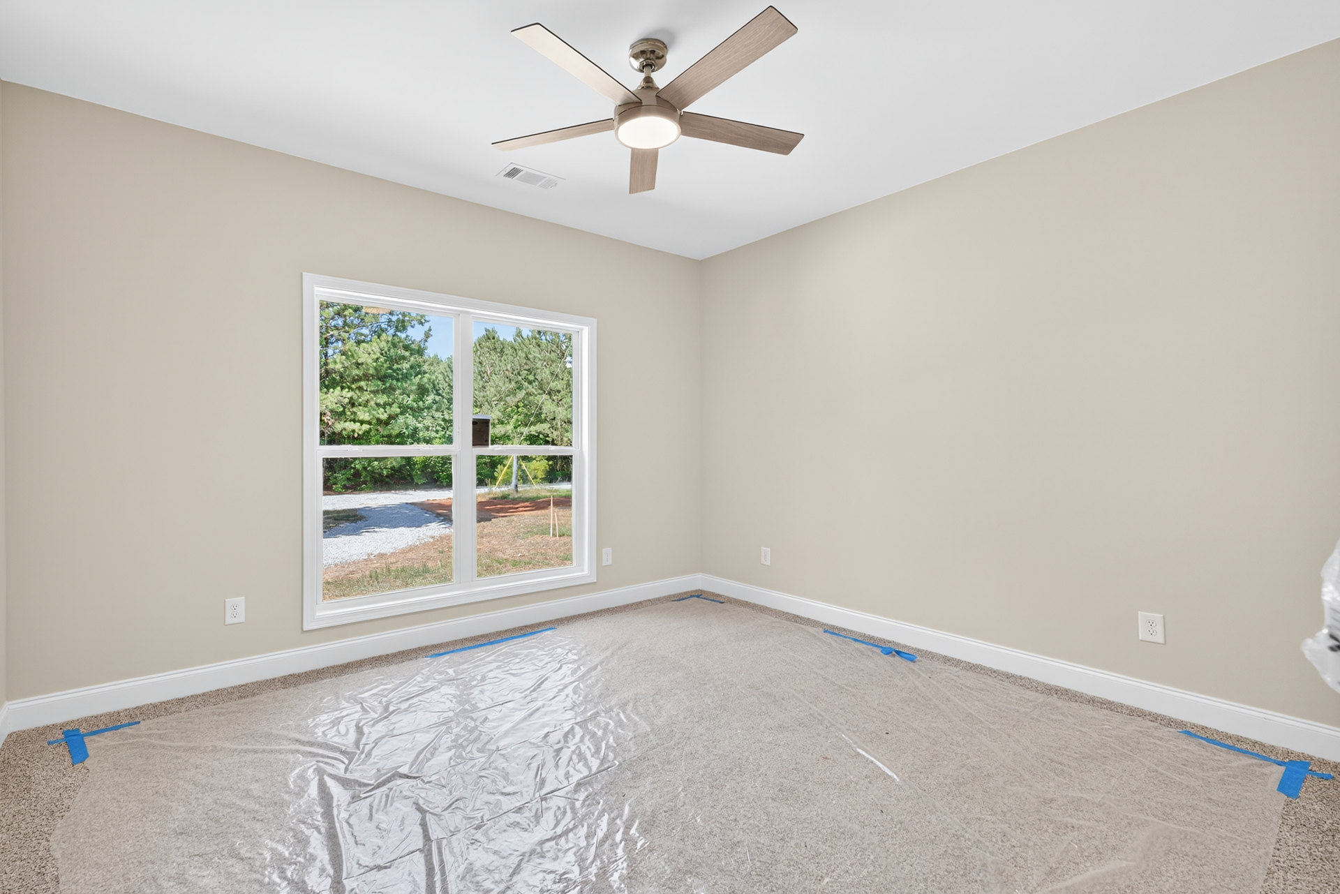Carpeted room with white walls, ceiling fan with light fixture, large window showing trees outside, plastic wrap covering part of the floor