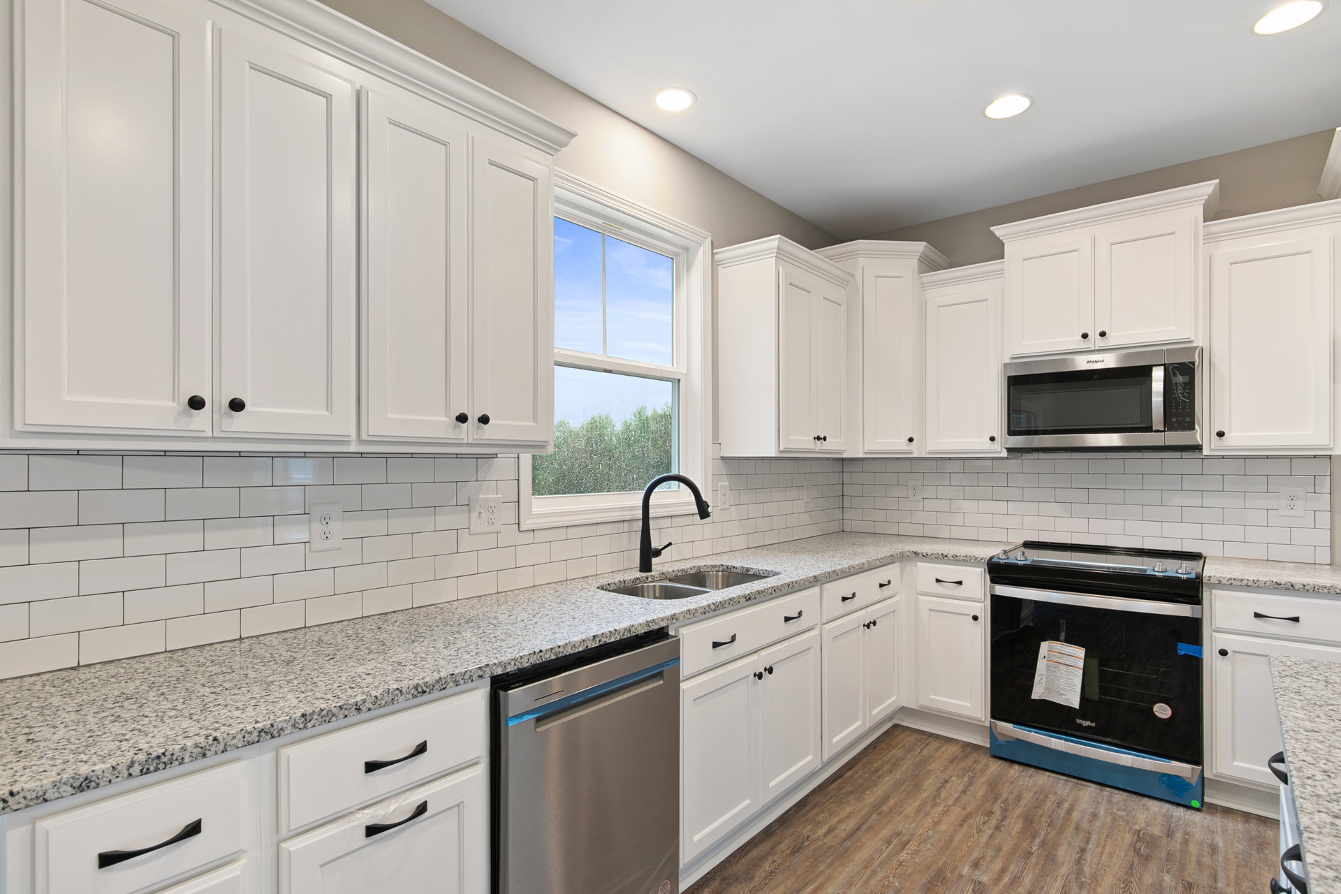 White kitchen cabinets, stainless steel dishwasher, black and silver stove, built-in microwave, window overlooking trees, light countertops