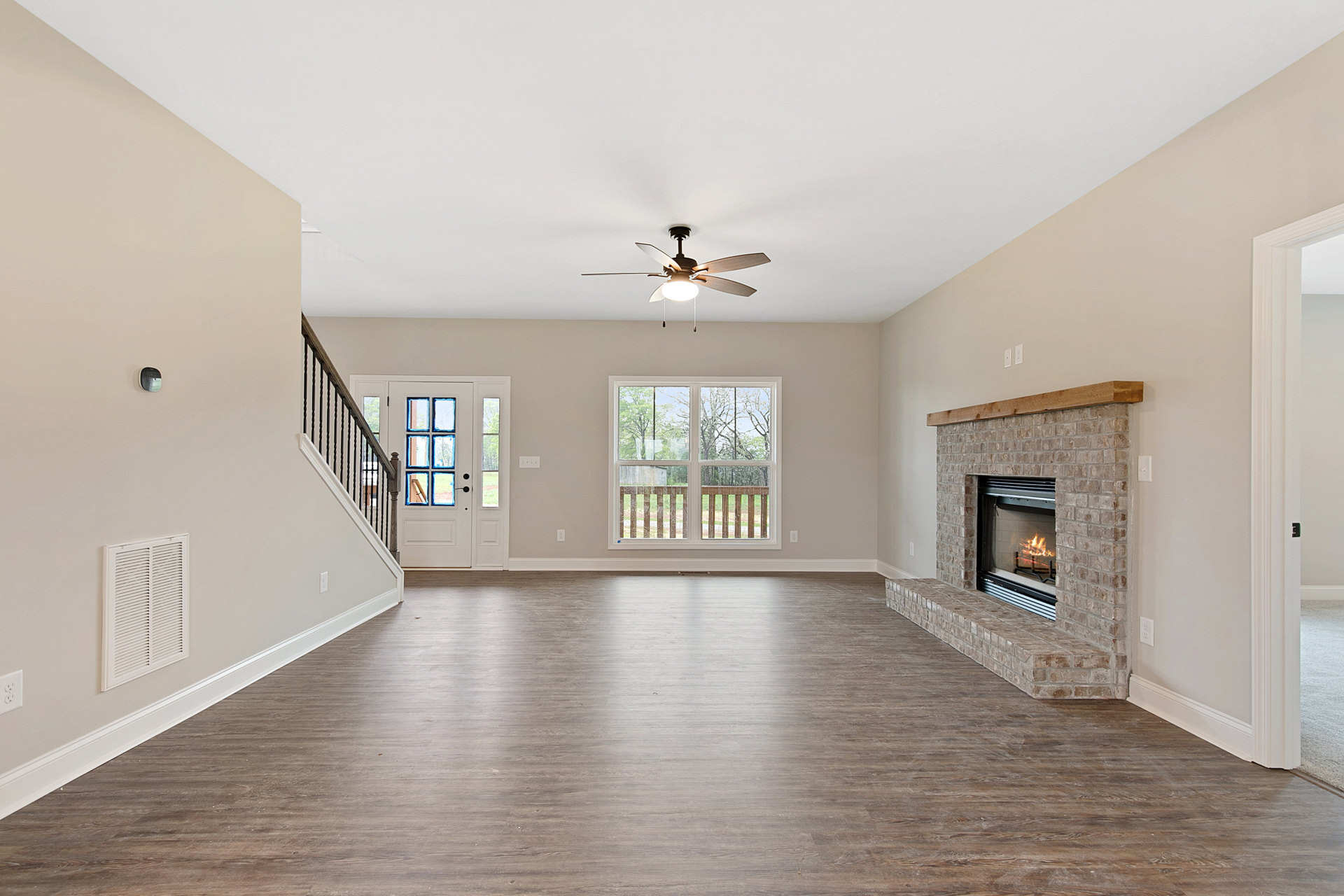 Living room with wood flooring, white plaster walls, stone fireplace with active fire, ceiling fan with light fixture, large window overlooking deck and trees.