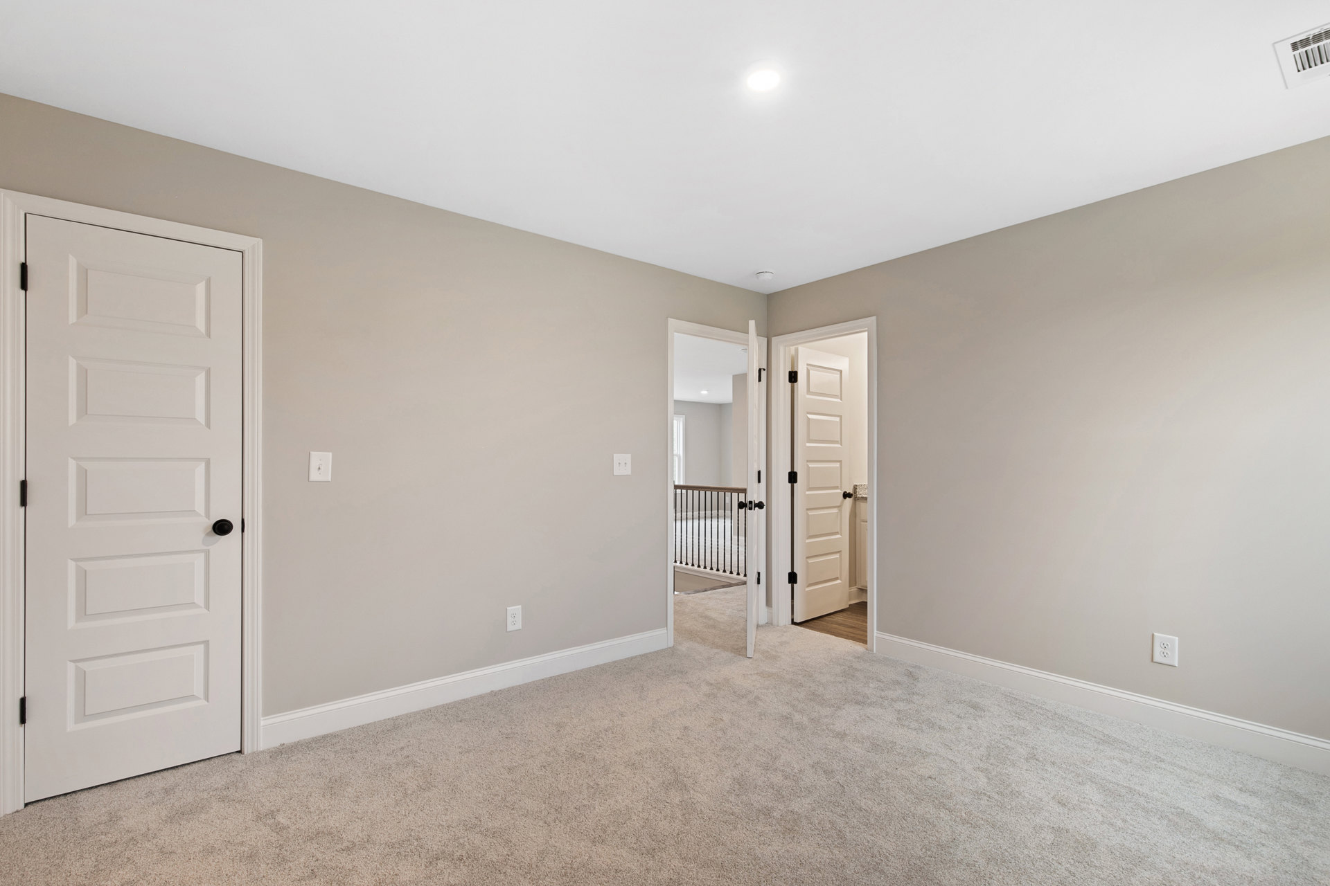 White-walled room with light gray carpet, open white door featuring black handle, ceiling light fixture, and partial view of stair railing
