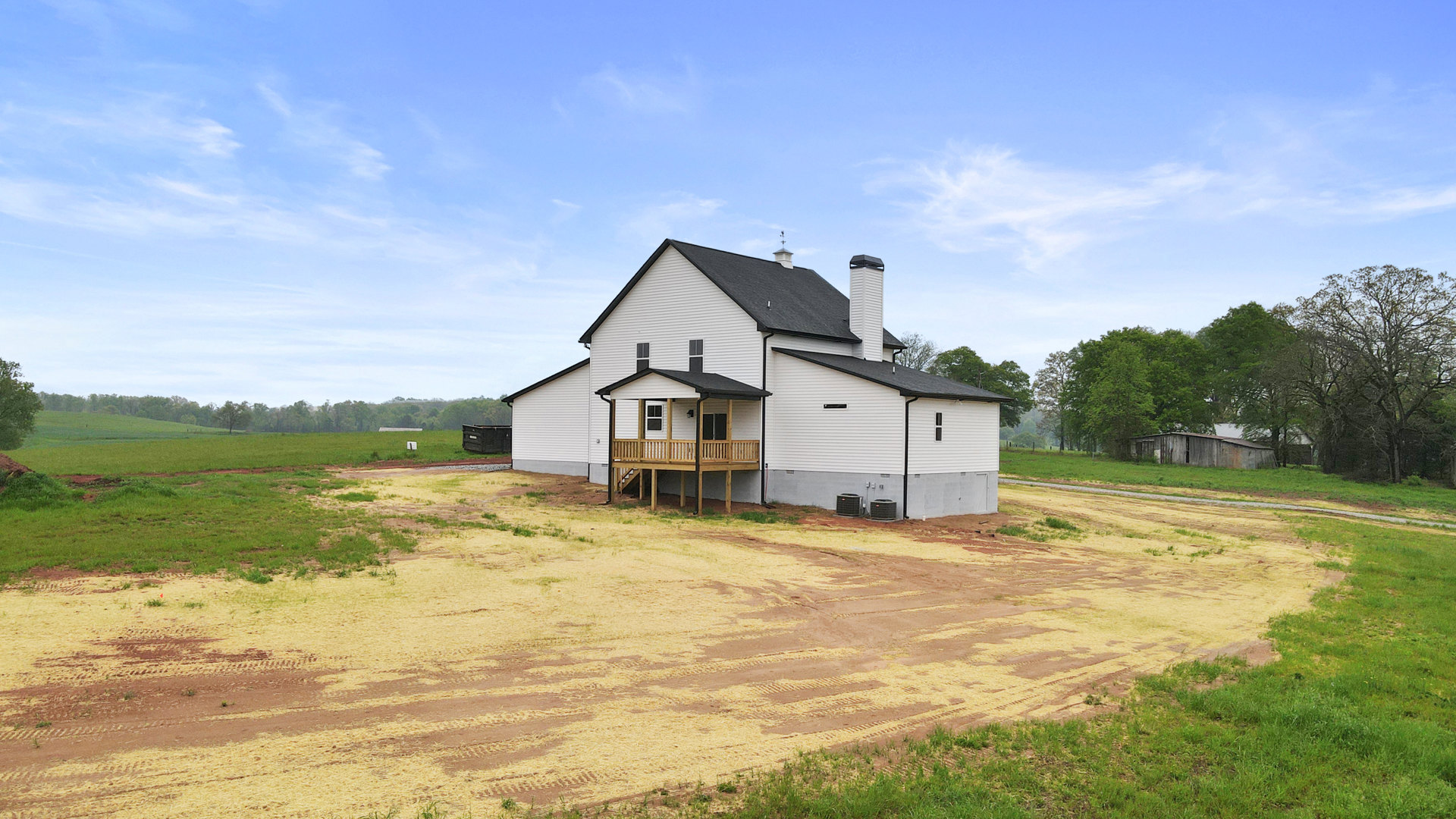 White house with black roof and chimney, wooden deck, spacious dirt yard bordered by grass and trees, partly cloudy sky overhead