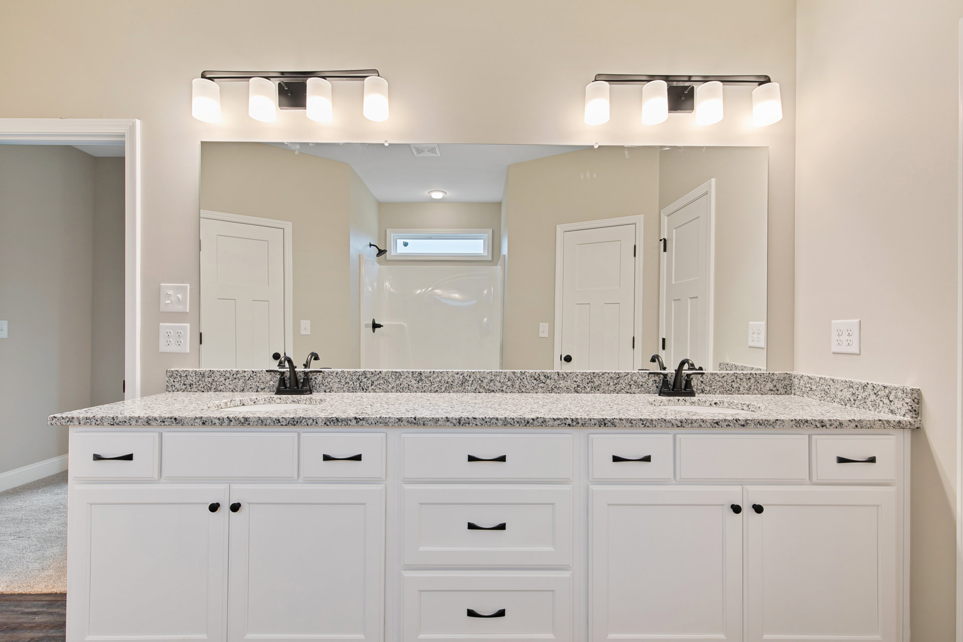 Bathroom with a wide mirror above a white vanity, black faucet and handles, tiled wall, visible electrical outlet and light switch, adjacent white door with black handle, partial