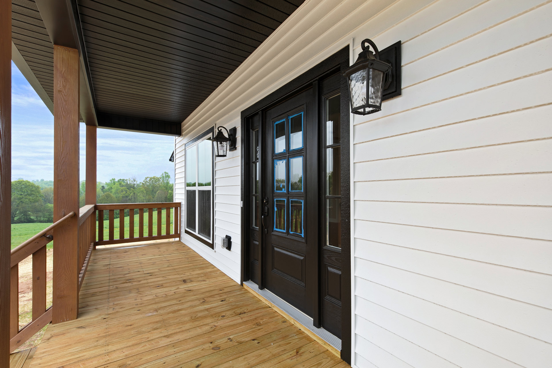 White siding house with black front door, wood deck and railing, porch lamp, and grassy field in background