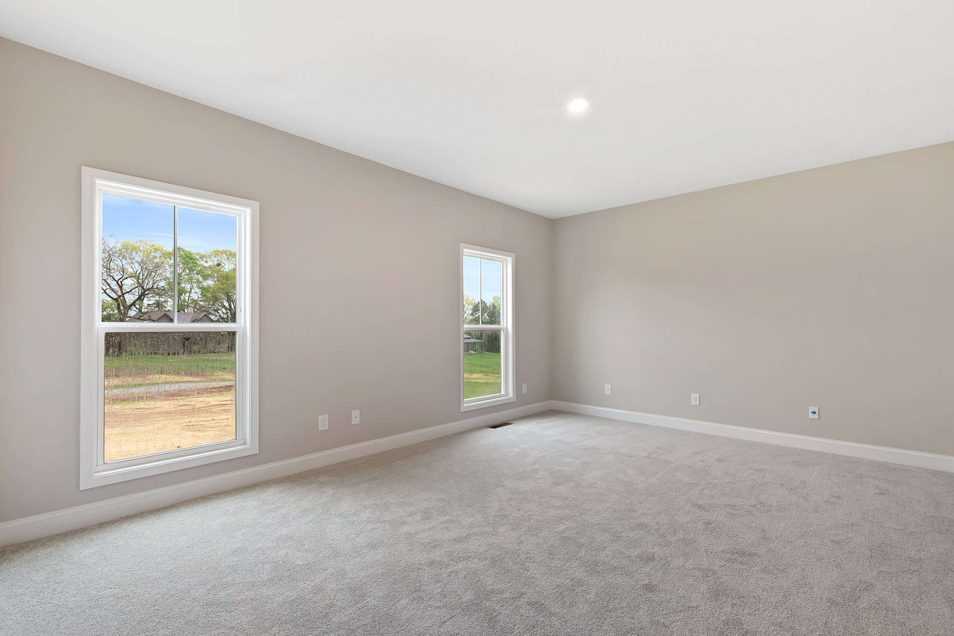 Bright carpeted room with large windows, white plaster walls, and ceiling; windows overlook a yard with trees and neighboring house.