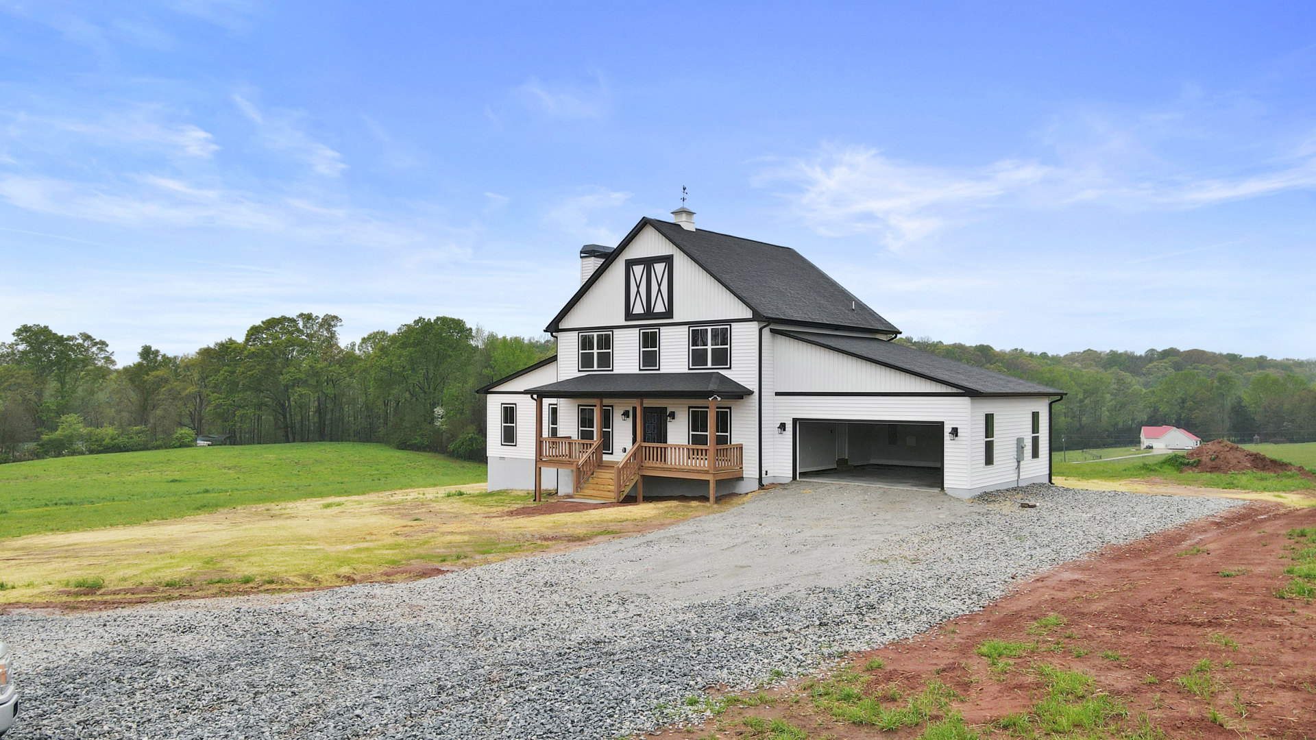 White house with black roof, attached garage, gravel driveway, wooden stairs leading to front entrance, wood deck, large windows with black curtains, surrounded by grass and trees