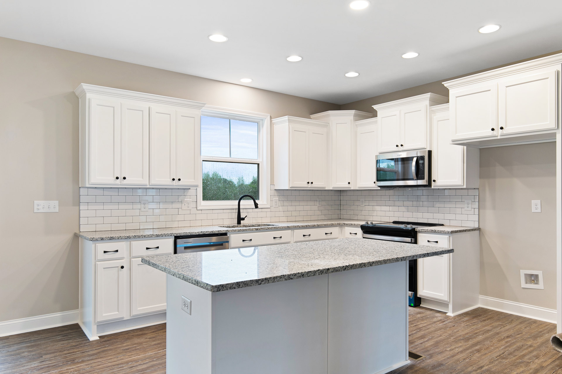 White kitchen with shaker cabinets, large central island with quartz countertop, black faucet at farmhouse sink, white subway tile backsplash, built-in microwave, window