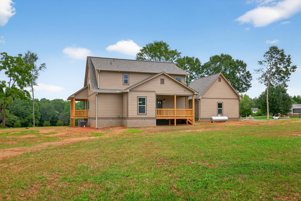White farmhouse with wood porch railing, manicured lawn, and tall leafy tree; blue sky and scattered clouds in background