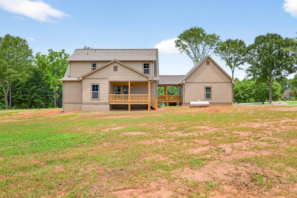 Two-story house with covered porch, wooden railing, and bench overlooking expansive grassy yard; mature tree with green leaves and partly cloudy sky in background