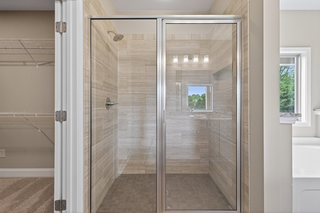 Glass shower enclosure with clear doors, tile flooring, and a window overlooking leafy trees
