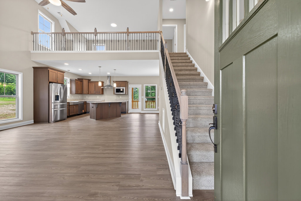 Open kitchen with wood flooring, stainless steel refrigerator, glass-paneled double doors, staircase featuring metal railing, large window overlooking grassy field and trees