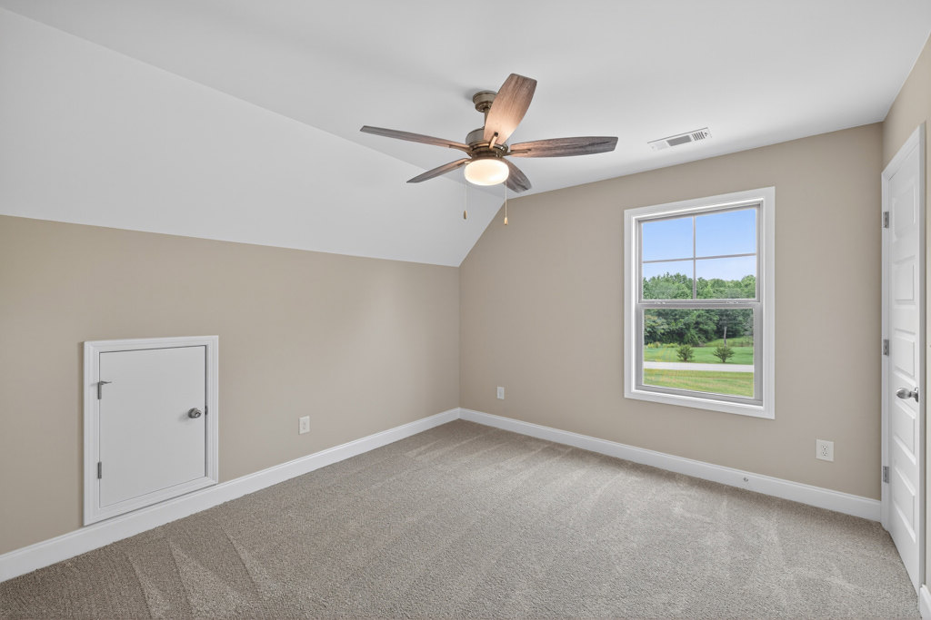 Ceiling fan with light fixture mounted on white plaster ceiling, carpeted room with white trim, window overlooking field and trees, white door with silver handle